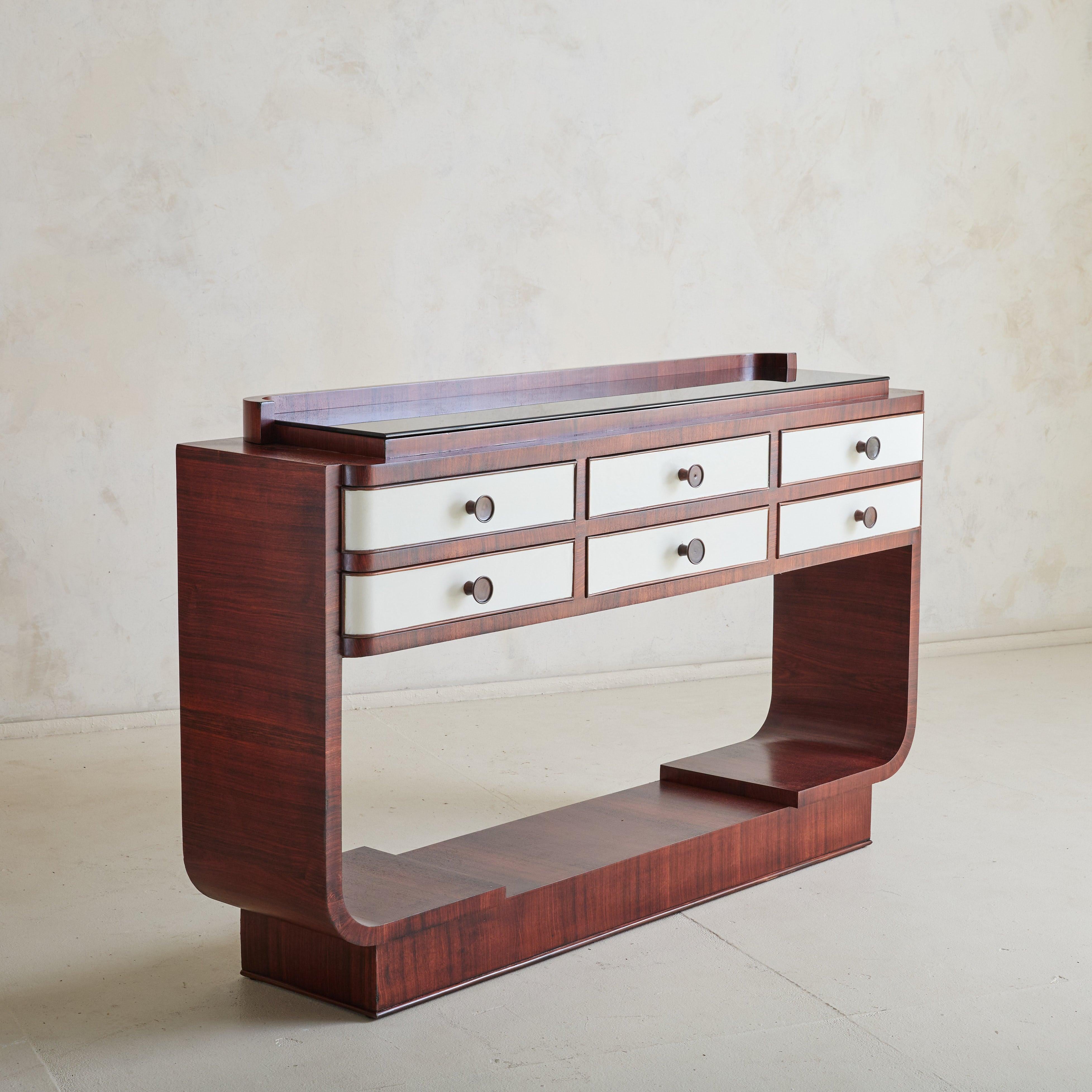 An Italian Art Deco console table from the 1940s, reminiscent of designs by Osvaldo Borsani. Crafted from rich rosewood veneer, this piece features six drawers covered in ivory vegan leather and accented by round wooden pulls. Its tiered surface is