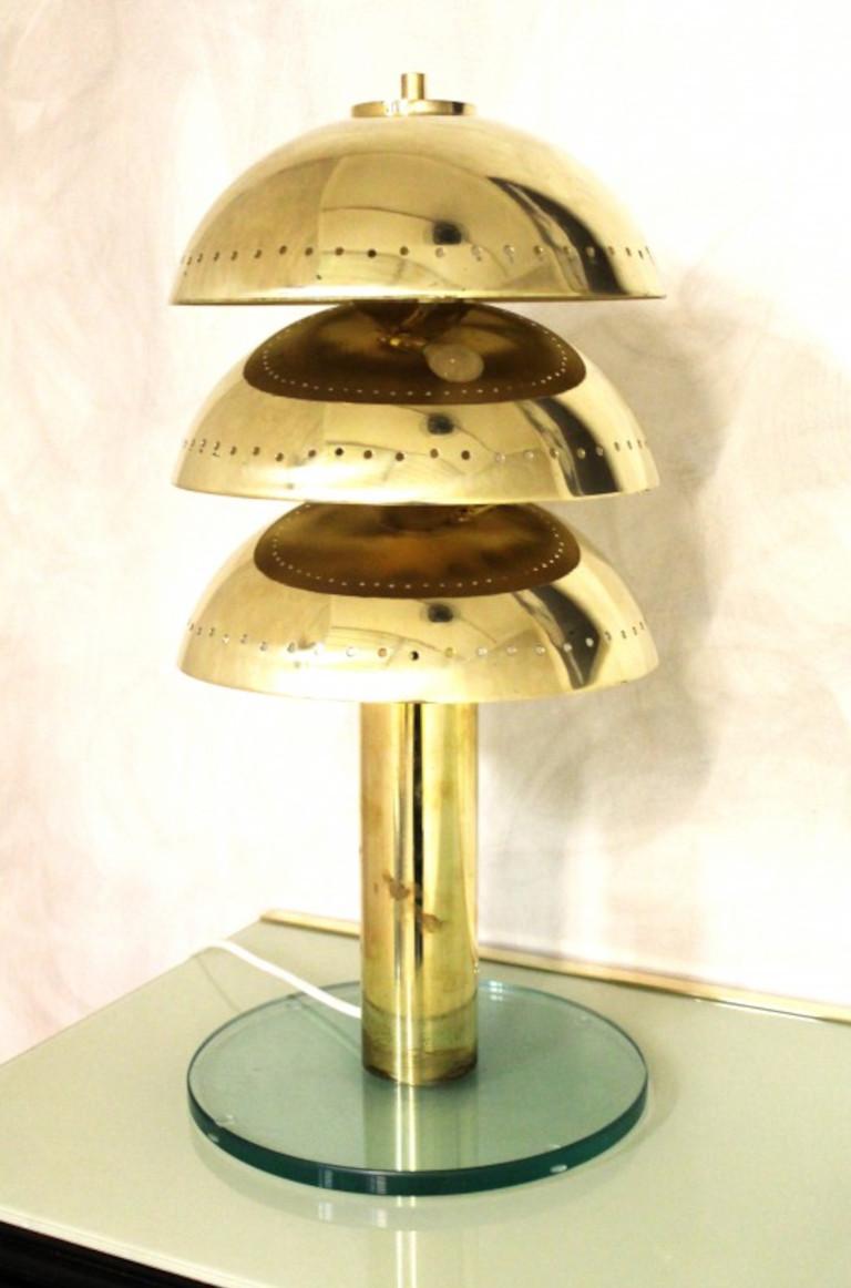 Lampada da tavolo in stile art déco italiano con tre livelli di paralumi in ottone traforato e corpo della lampada in ottone naturale non laccato, montato su una base in vetro trasparente smussato / Made in Italy
Altezza: 23,5 pollici / diametro: