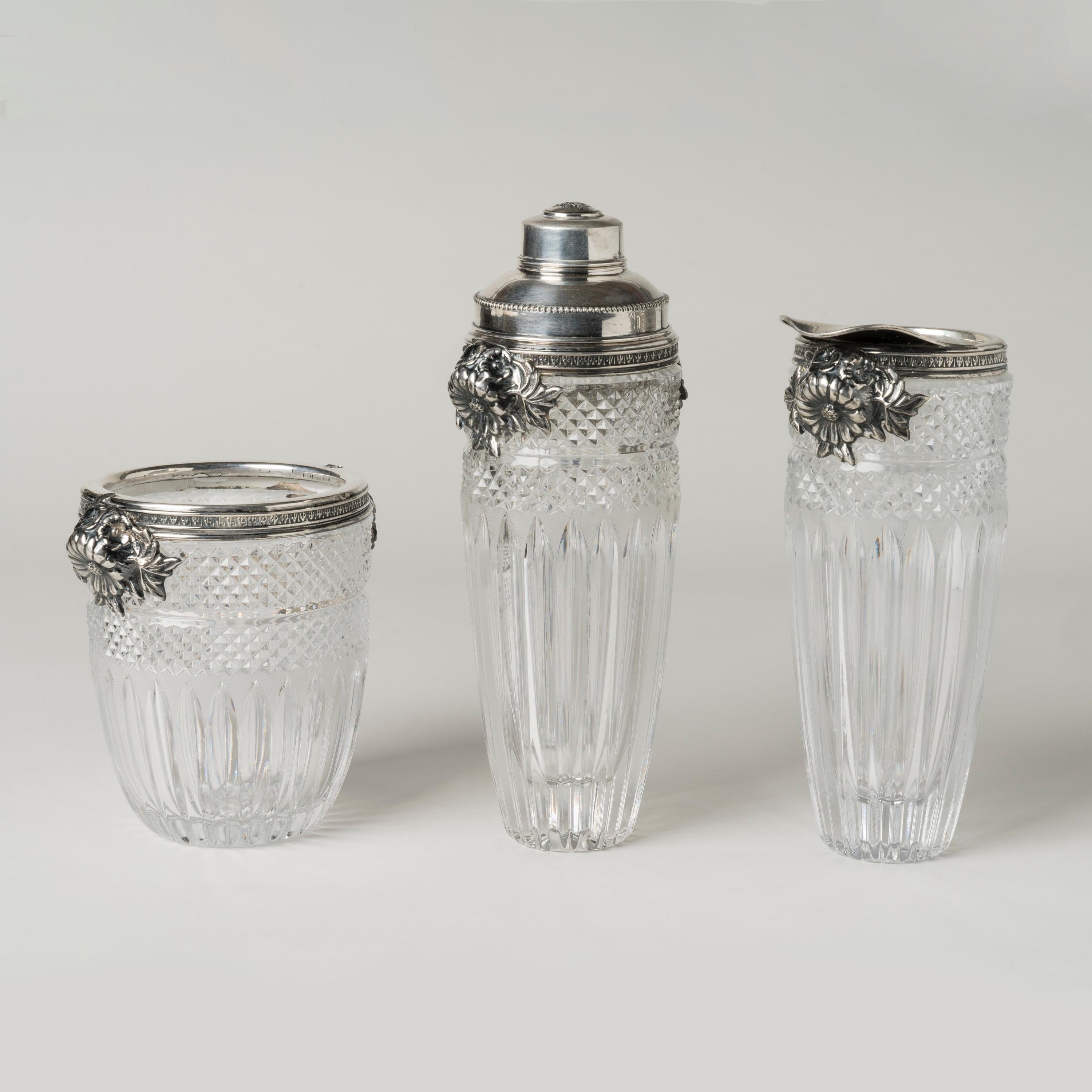 **Please ask for specific dimensions if needed**

Consisting of a shaker, ice bucket and pitcher, with hand-cut glass bodies of diamond cut pattern fading into a prism cut pattern, the silver collars around each consisting of flowers in bloom