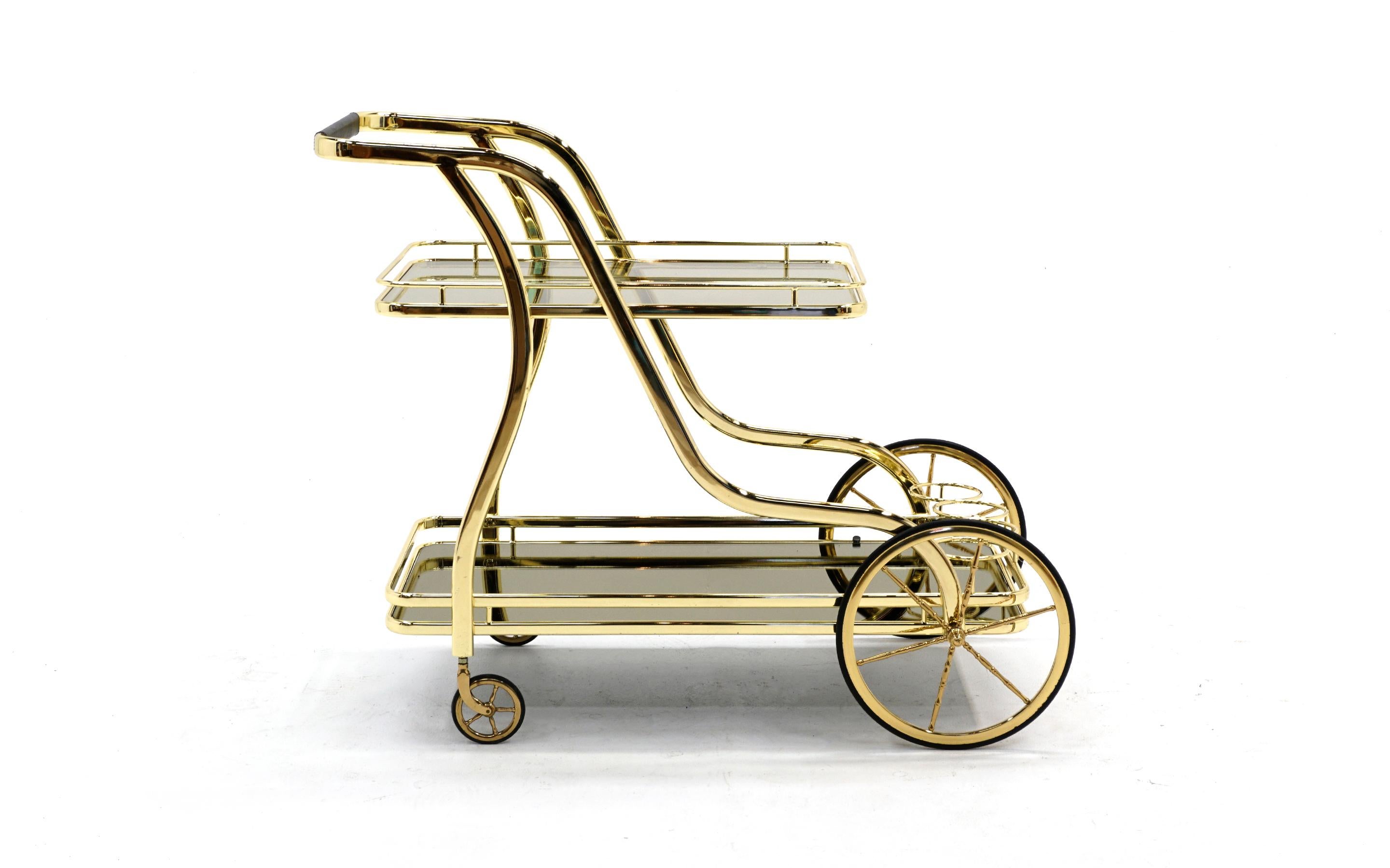 Serving / bar cart in brass with a clear glass top shelf and bronze mirrored glass lower shelf. This creates striking visual effect. Large front wheels with rubber tires and small rear wheels. Ready to use.