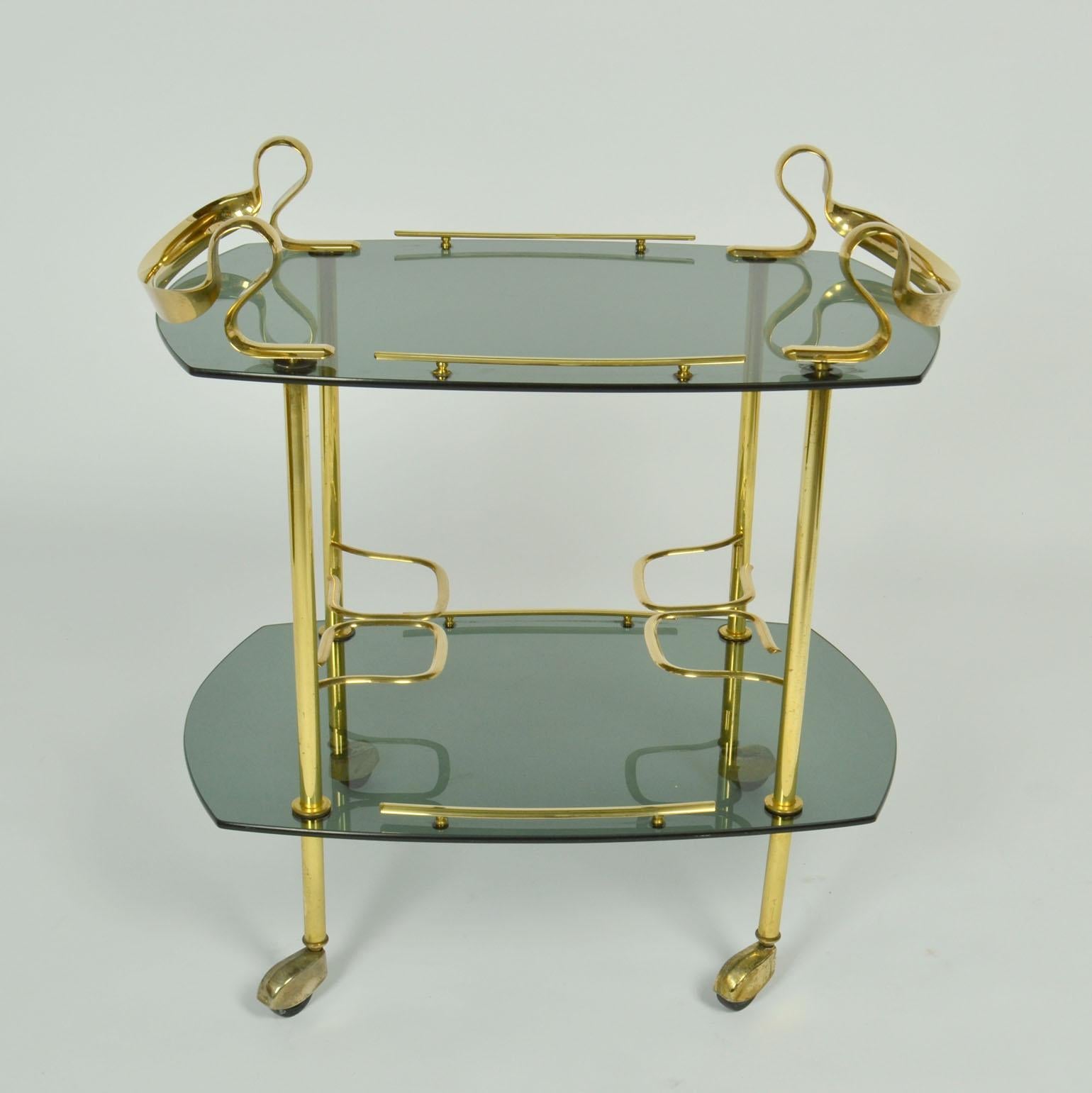 This is an unusual elegant bar cart, cocktail or serving trolley in brass and smoked glass Italy circa 1960's. The sculptural  design celebrates social gatherings with the curvaceous serpentine brass handles and arched cut glass trays. 