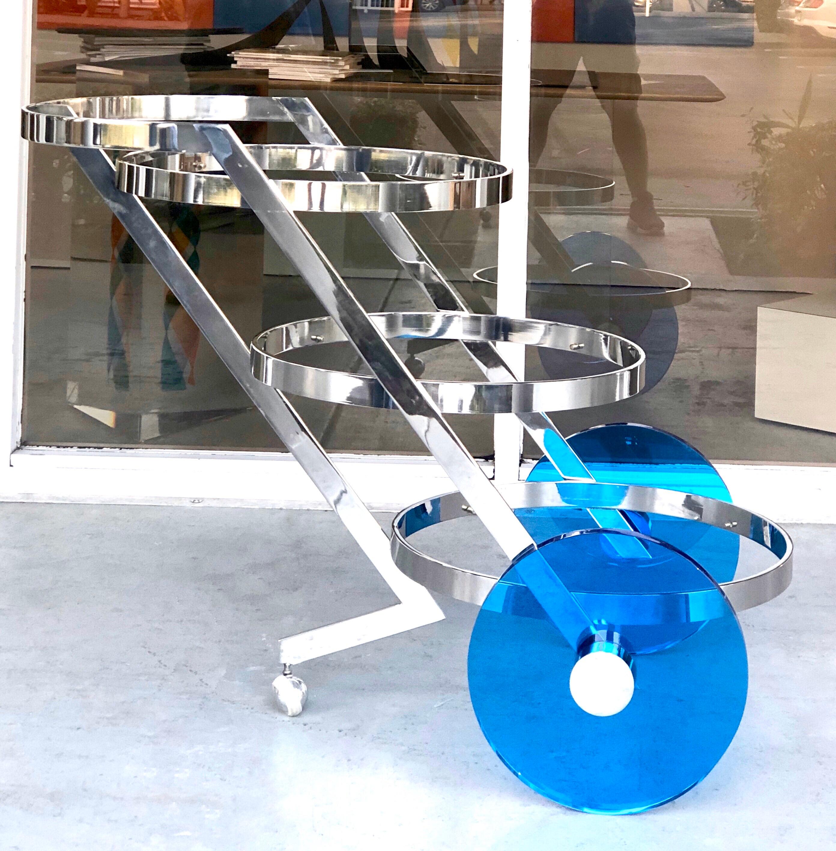 A striking stainless steel bar cart with blue Lucite wheels. 3 round cantilevered glass shelves.