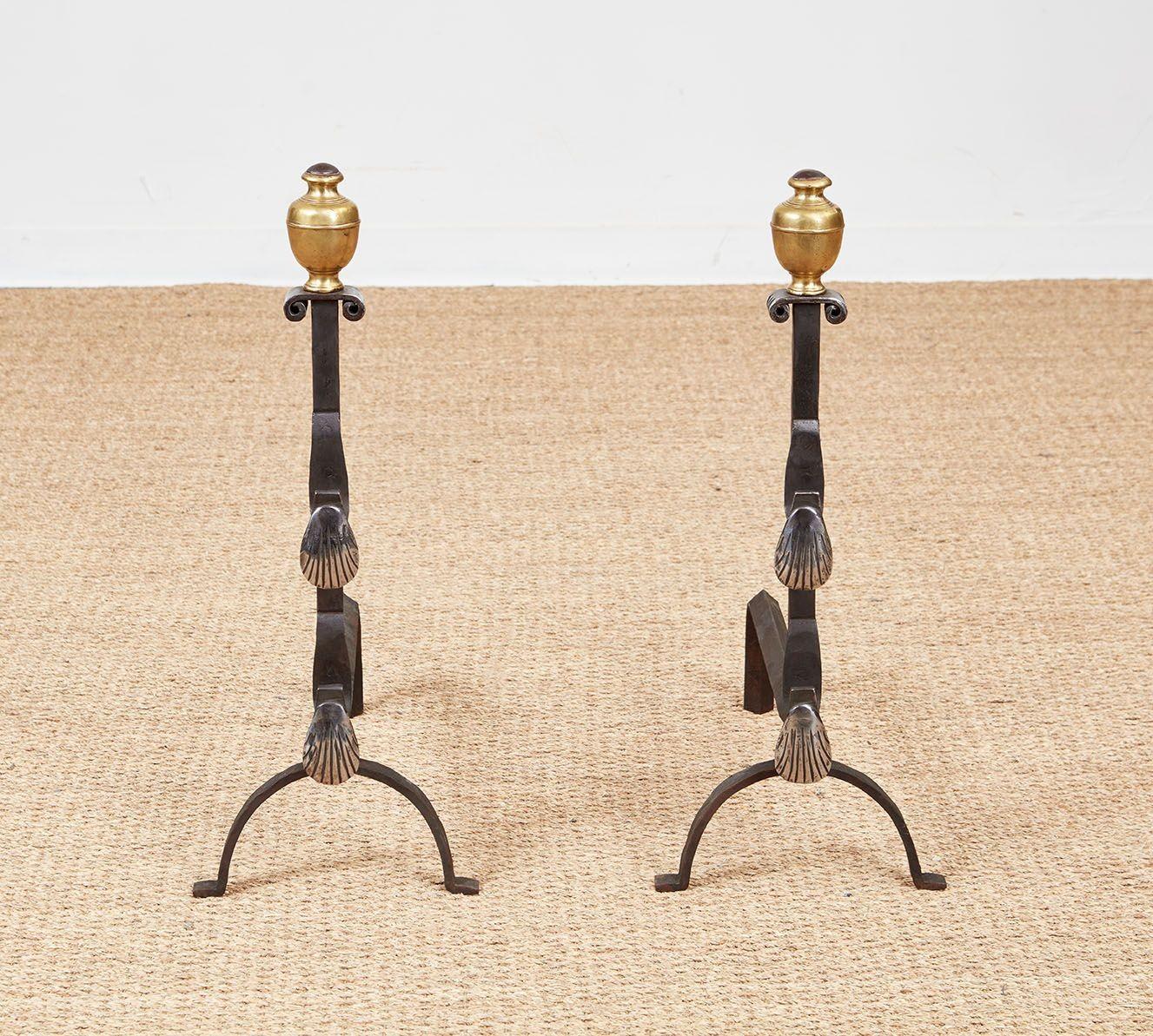 A pair of andirons with double scallop shell spit holders of polished iron on black iron bodies and arched legs with penny feet, topped by decorative urn shaped brass finials sitting on a scrolled iron platform at the top of the shaft.  Italian