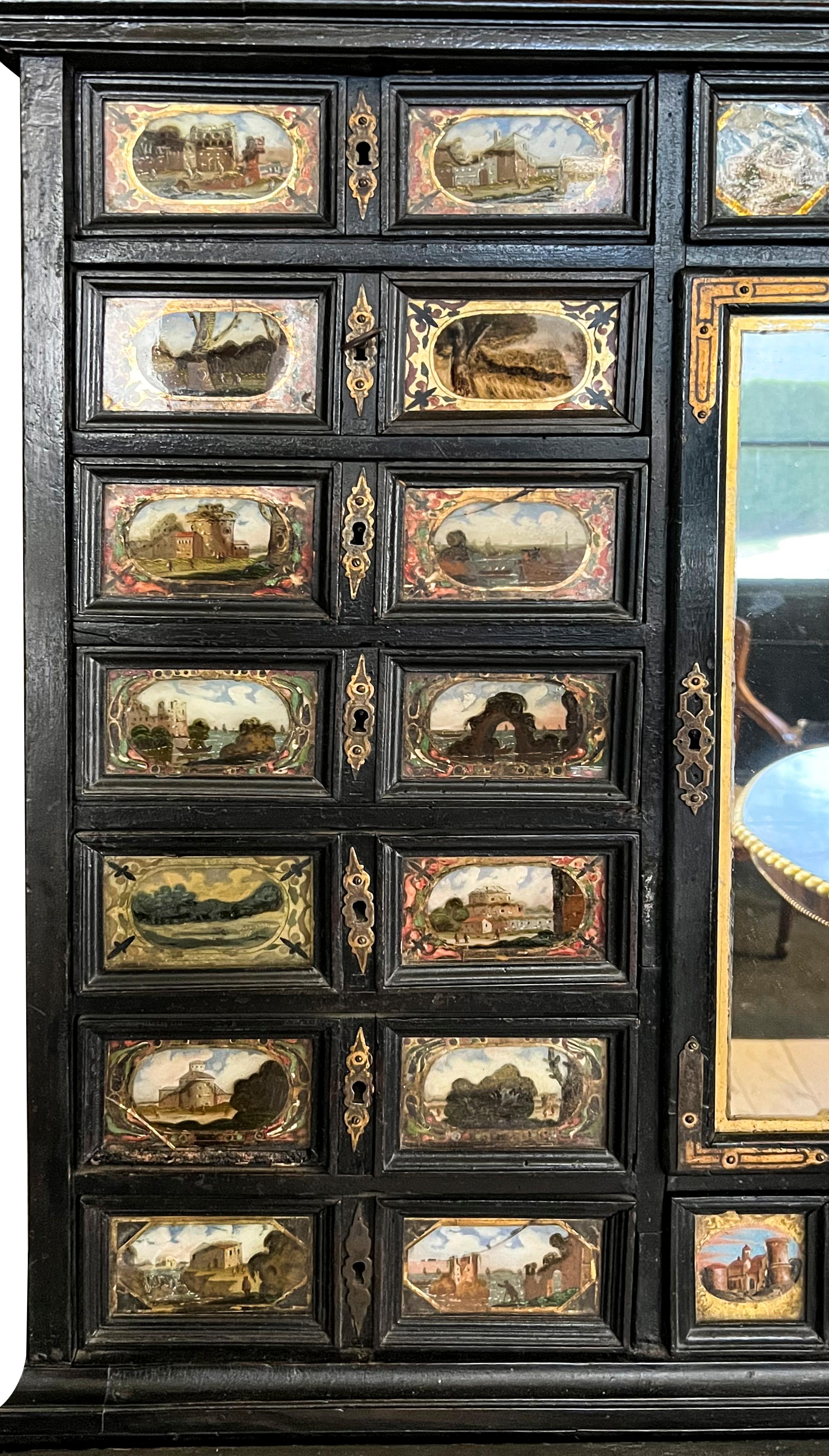 Slightly arched top that may have held a bust at some point, over a middle section with drawers with reverse painted panels with scenes of ancient towns and ruins with a central mirrored door, the base with hinged slant lid opening to a fitted