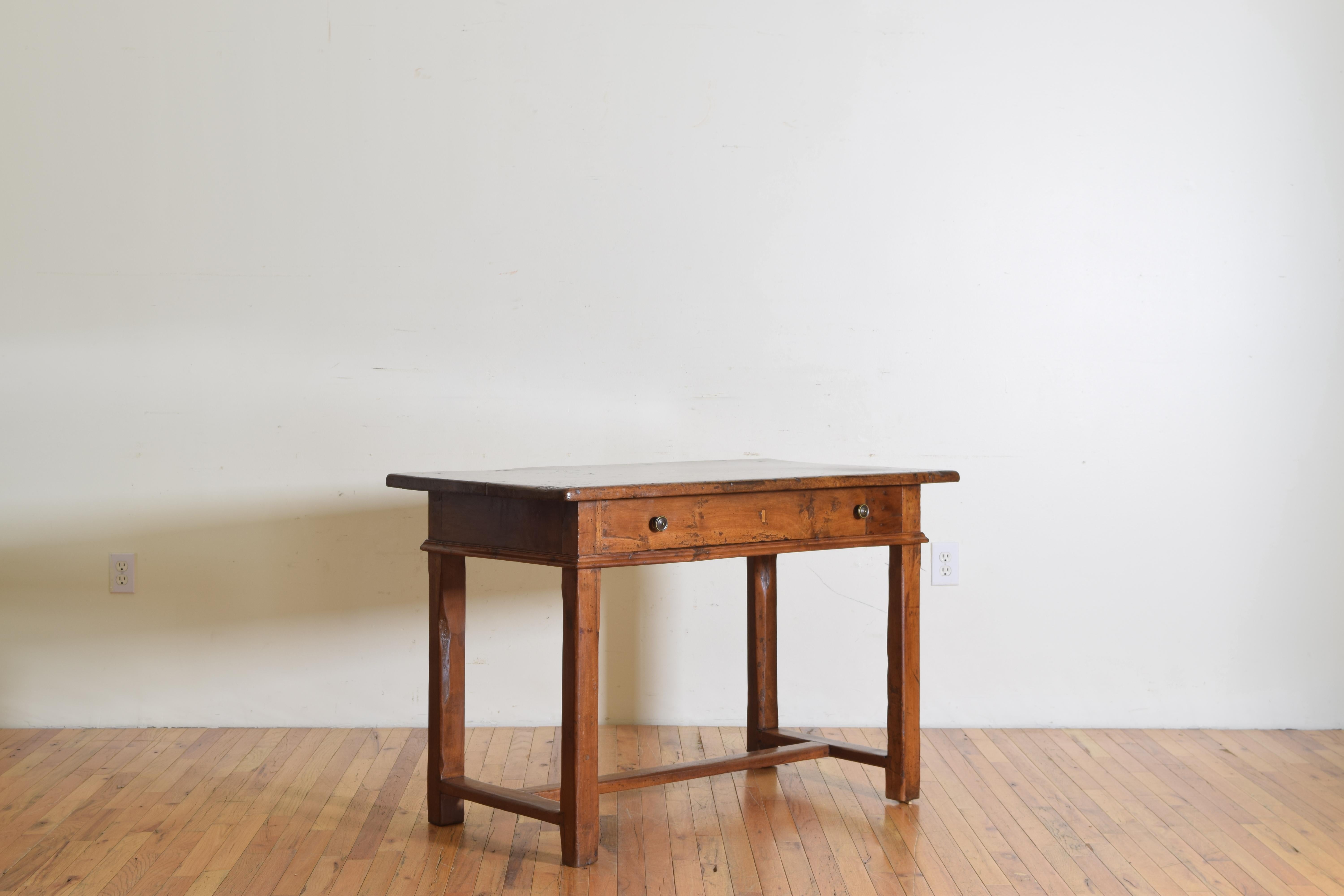 Having a thick solid walnut top overhanging a case with a lower molded edge and housing a single long drawer with two bronze pulls, raised on block legs joined by an H-form stretcher.