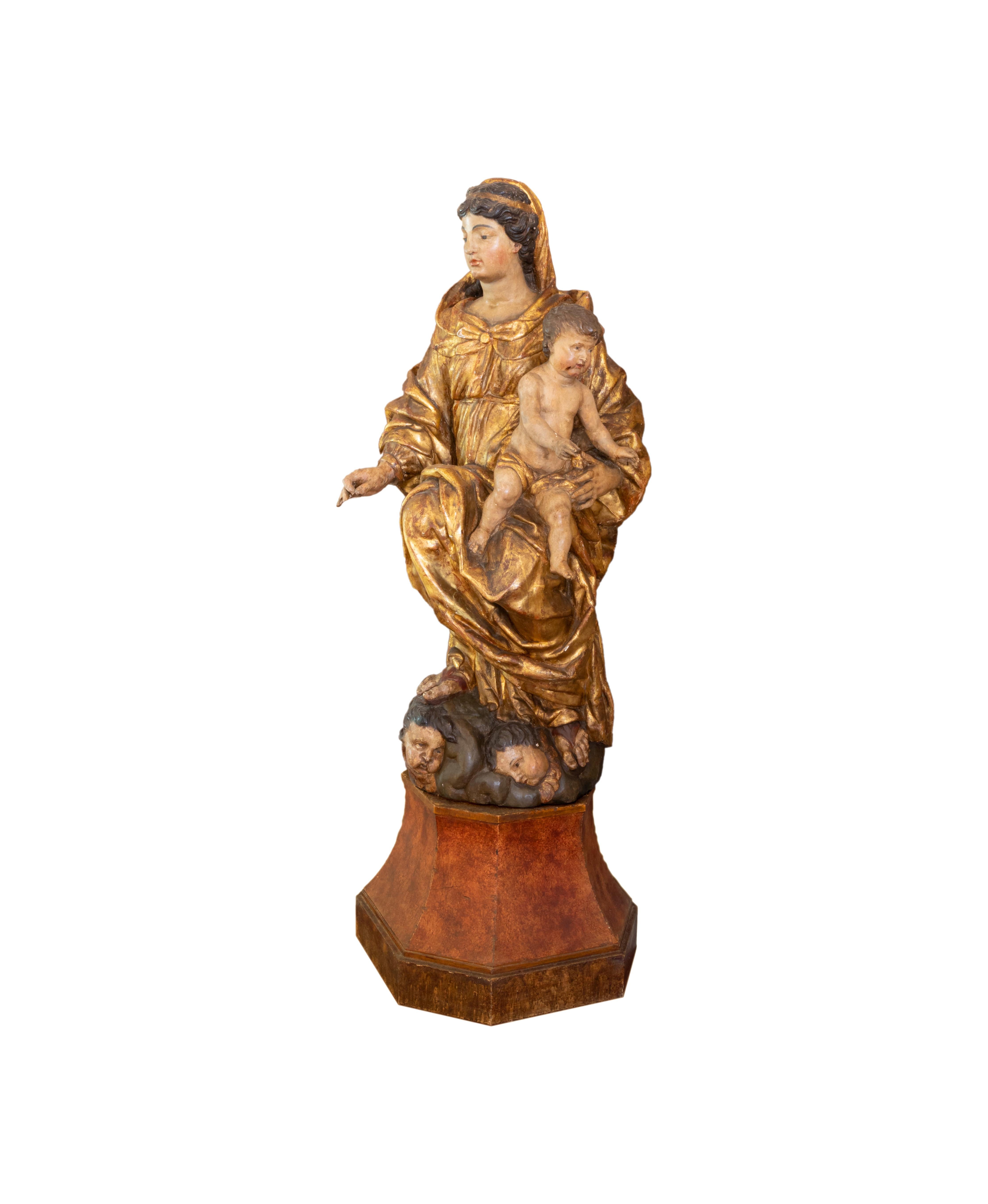 A 18th Century superb italian sculpture of a draped Madonna with a veil, base with richly carved relief with cherubs and a baby Jesus on her lap in carved polychrome and gilded wood. The scale, presence and skillful built of the sculpture impose the