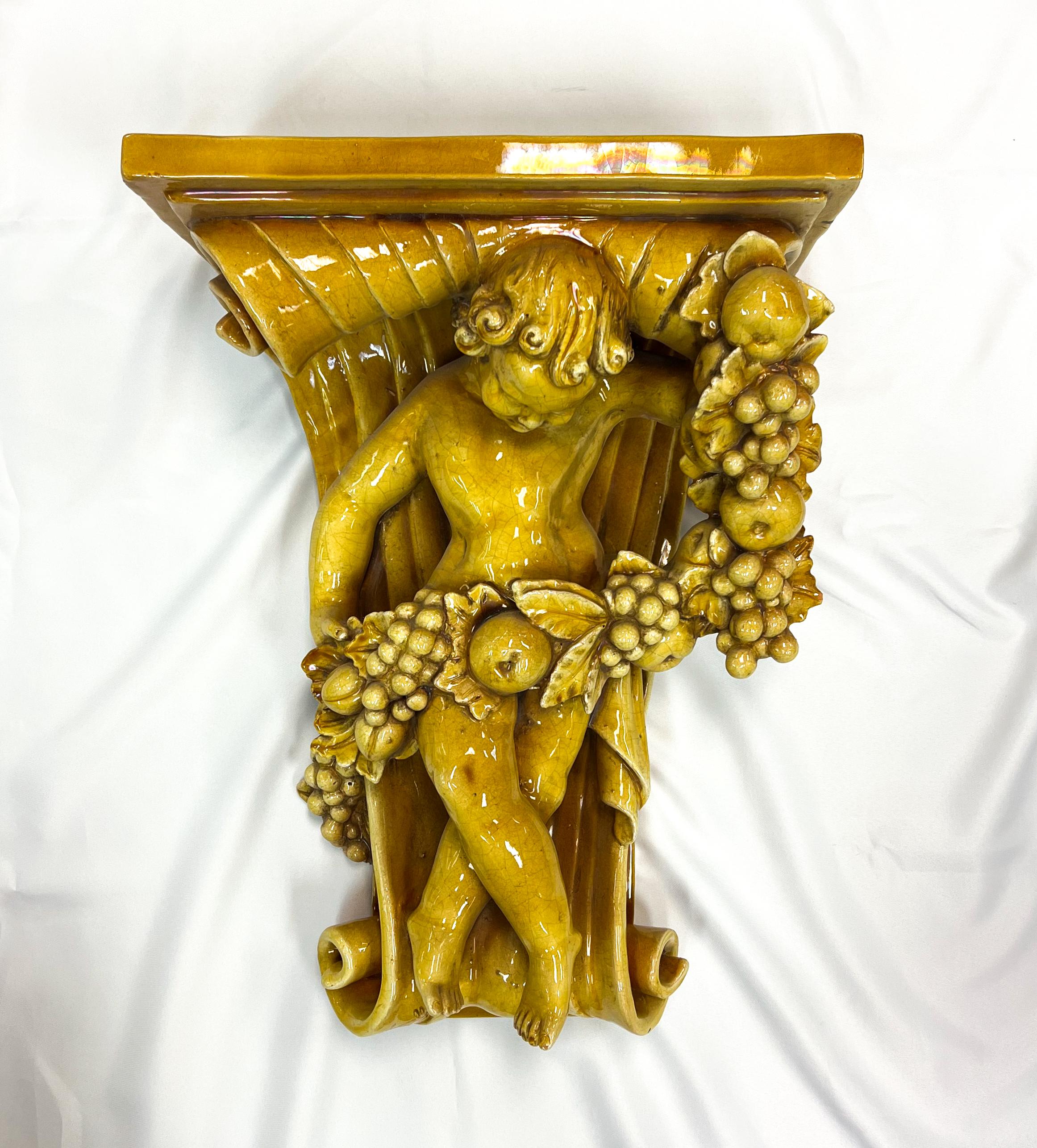 A handcrafted Italian glazed pottery putto wall bracket from the 1920s in mustard color and intricate details.

The putto, a cherubic figure holding a vine of fruits, showcases the artistry and craftsmanship of the Italian potter. The choice of