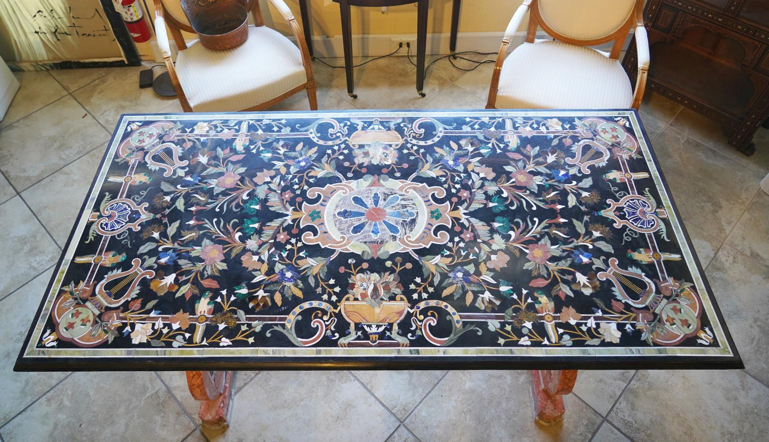 Measuring 36 x 72 inches and dating to the 20th century this grand Pietra Dura dining table features an elaborately inlaid stone top with flowers, classical themes and cartouches in a multitude of colors. The table top rests on twin carved and