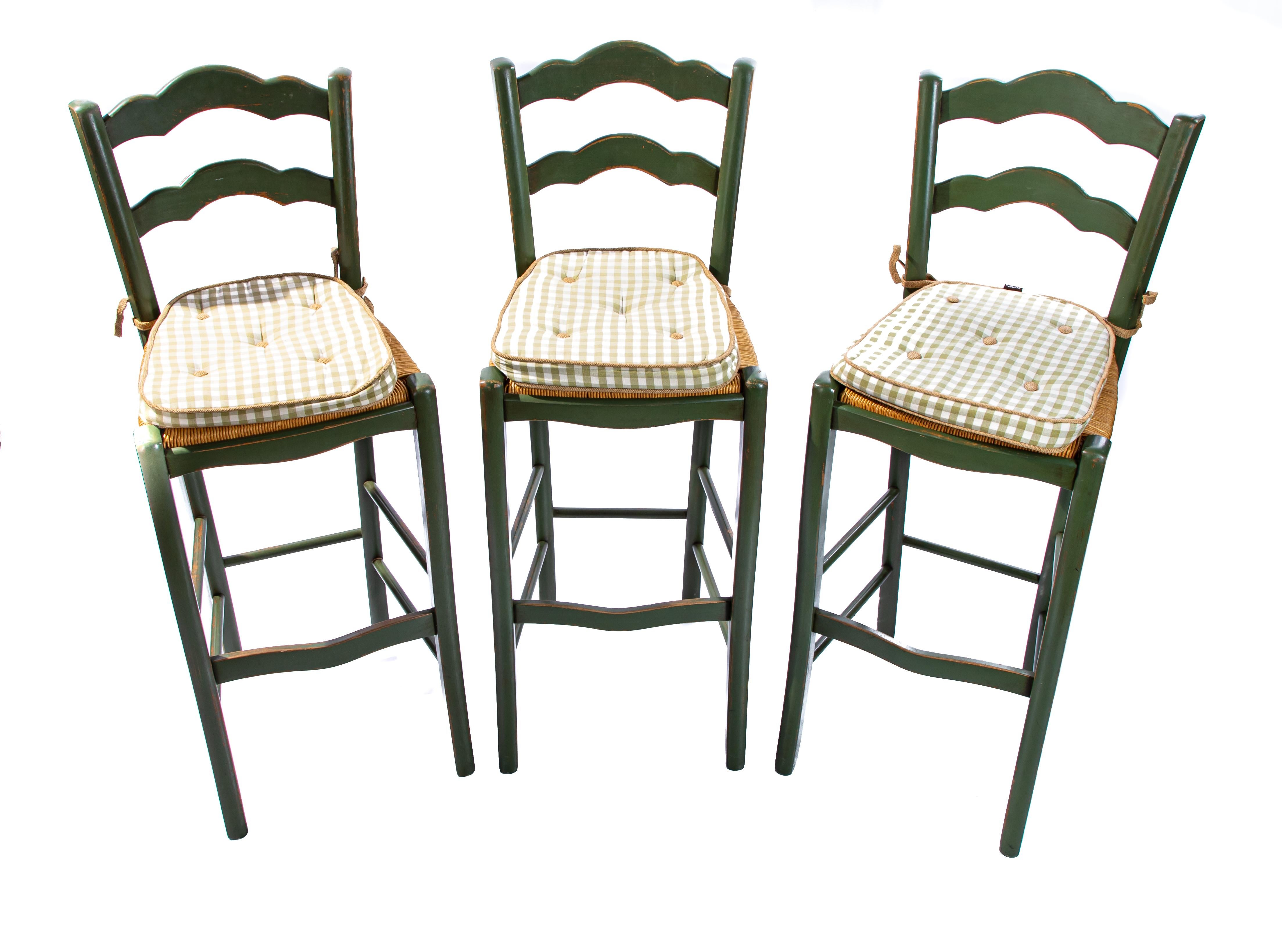 Offering three beautiful Italian barstools painted in green. Sitting atop a simple cabriole leg and curvy stretchers. They have rush seats and plaid upholstered pads. They all three have ladder backs. Marked on the underside, made in Italy.