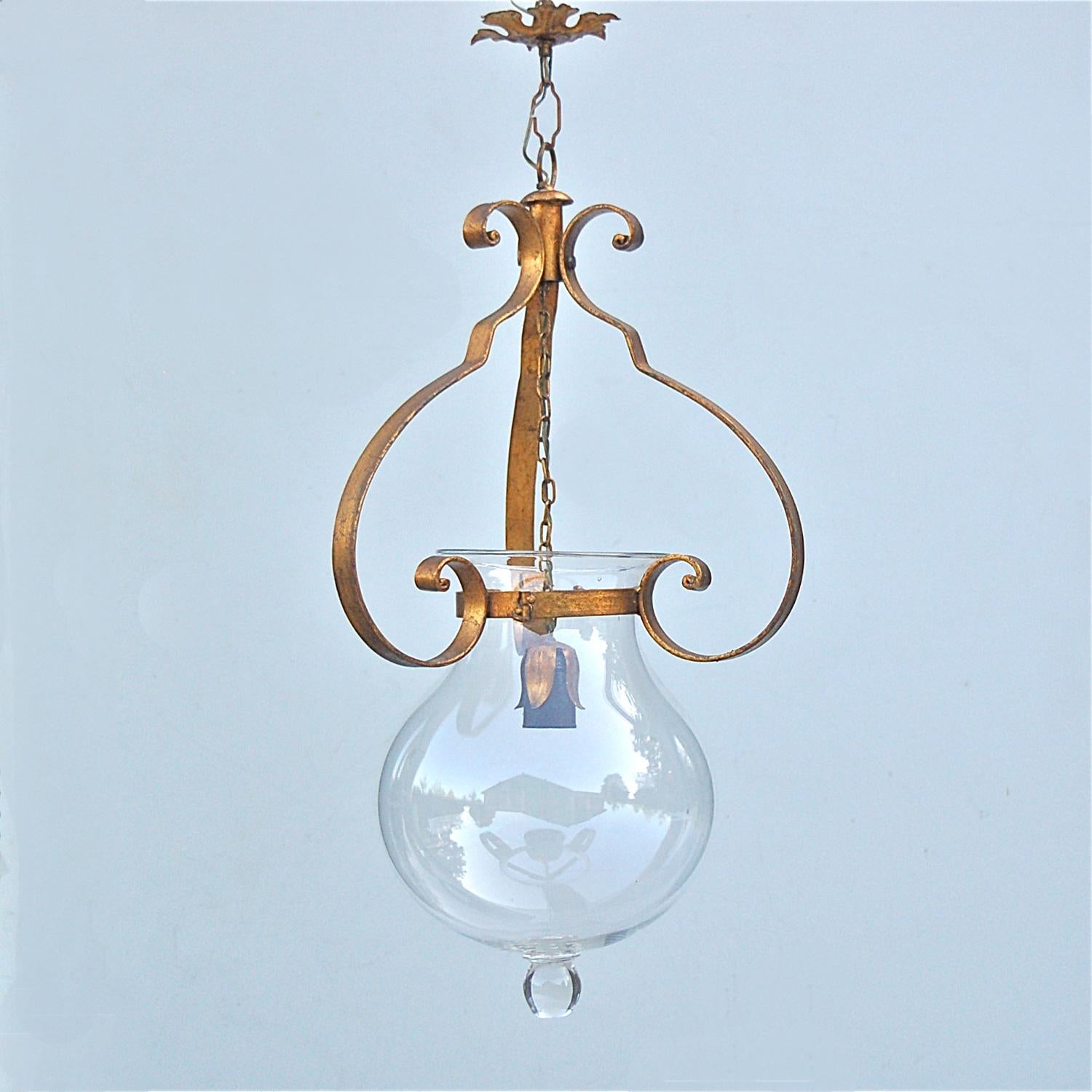 An Italian bell jar pendant or hallway lamp. The design is Inspired by the antique hall lantern. A thick, clear, bell jar shaped piece of glass sits in a metal ring attached to three broad, swirling, curved metal bands or arms with an antique gold