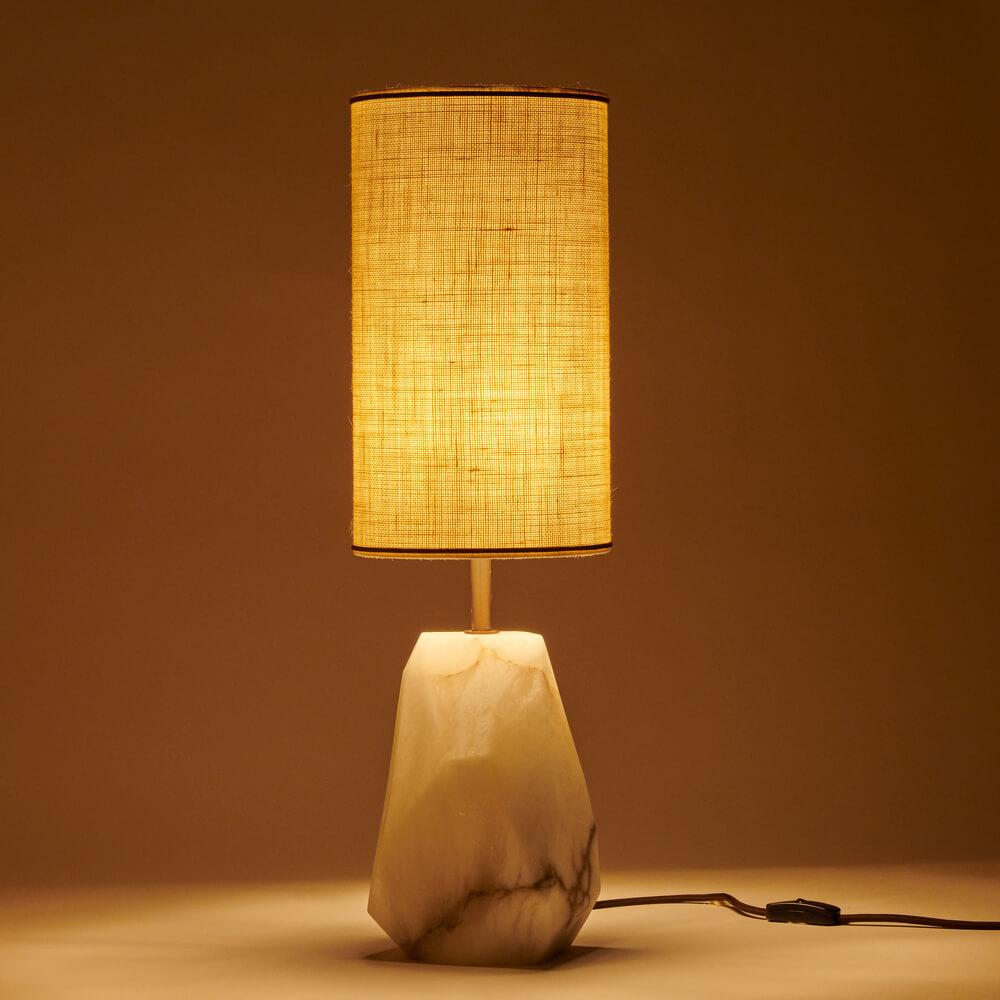 The Monolite table lamp is a stunning piece that combines elegance and sophistication with a touch of natural beauty. Its base is crafted from a raw block of alabaster that has been faceted, creating a visually appealing and unique design. Alabaster