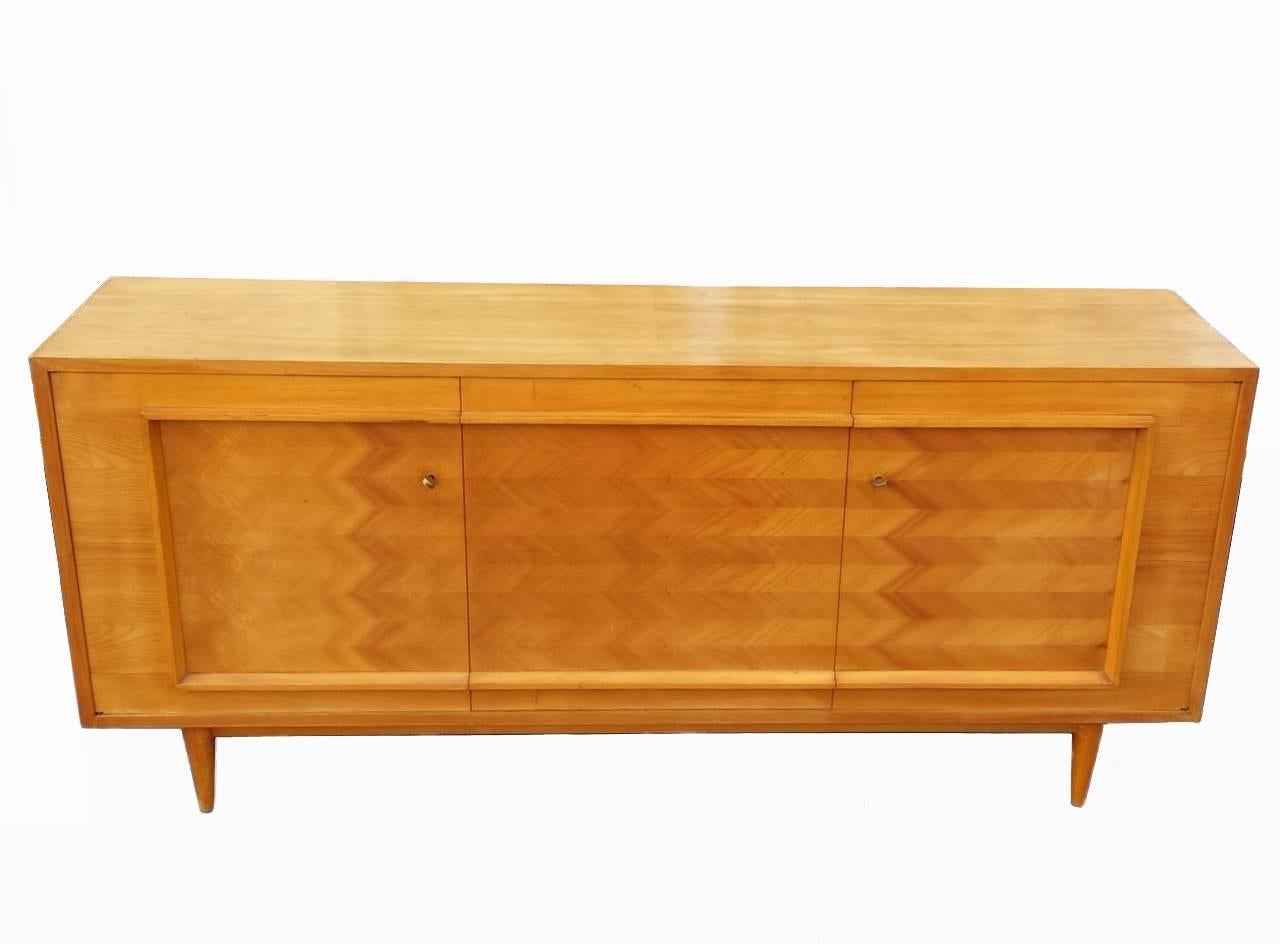 Blond cherry veneered three door credenza with a framed fish bone pattern front. The credenza has cutlery drawers and  shelves behind the outer doors. The middle door comes down to allow a bar or large equipment. There are two original brass keys