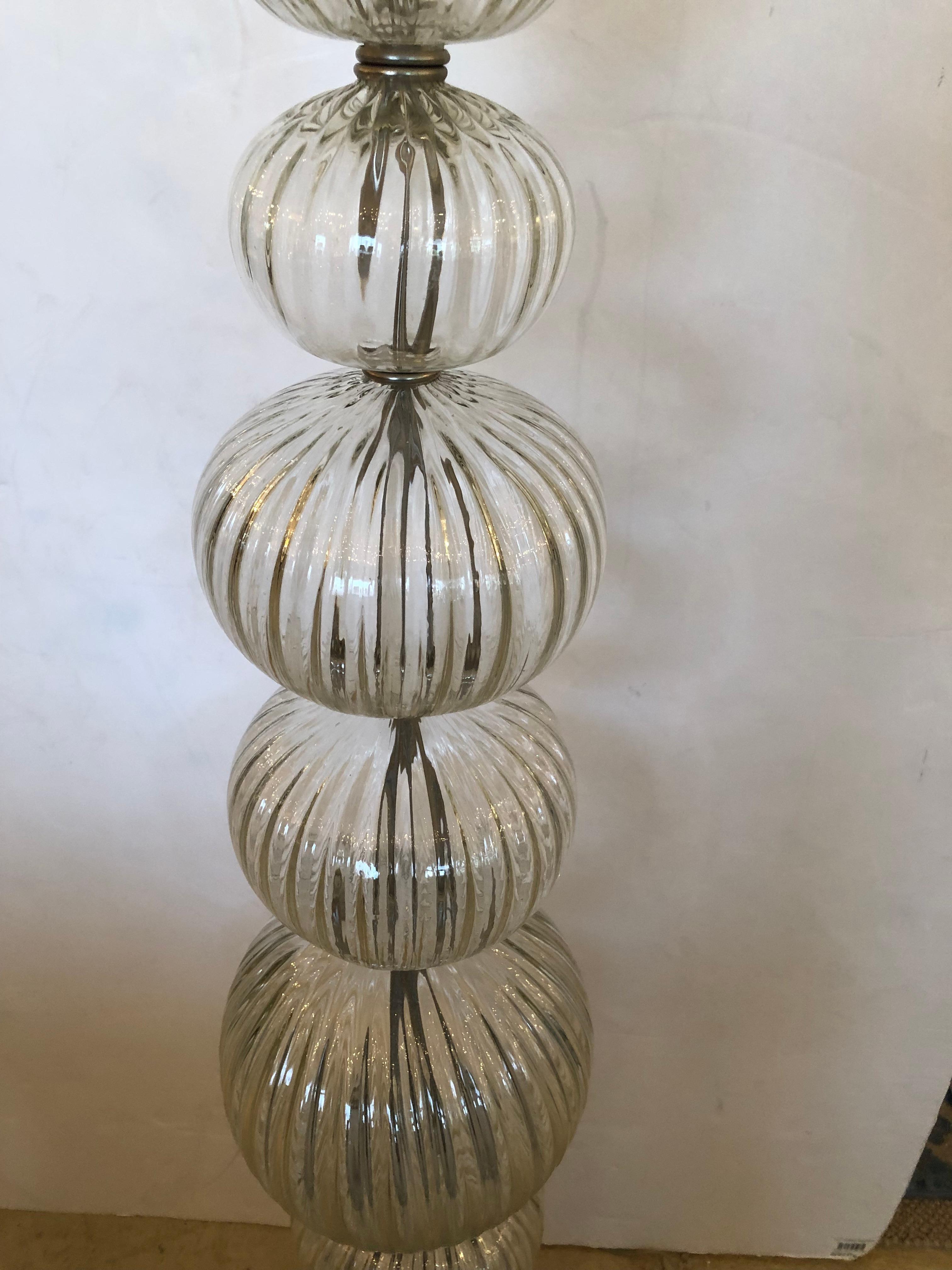 Elegant lampadaire en verre transparent de Murano avec une colonne composée de globes ronds nervurés empilés avec de l'acier entre les deux et une base ronde en acier.  Magnifique fleuron en bronze en forme de flamme au sommet.
L'ombrage est
