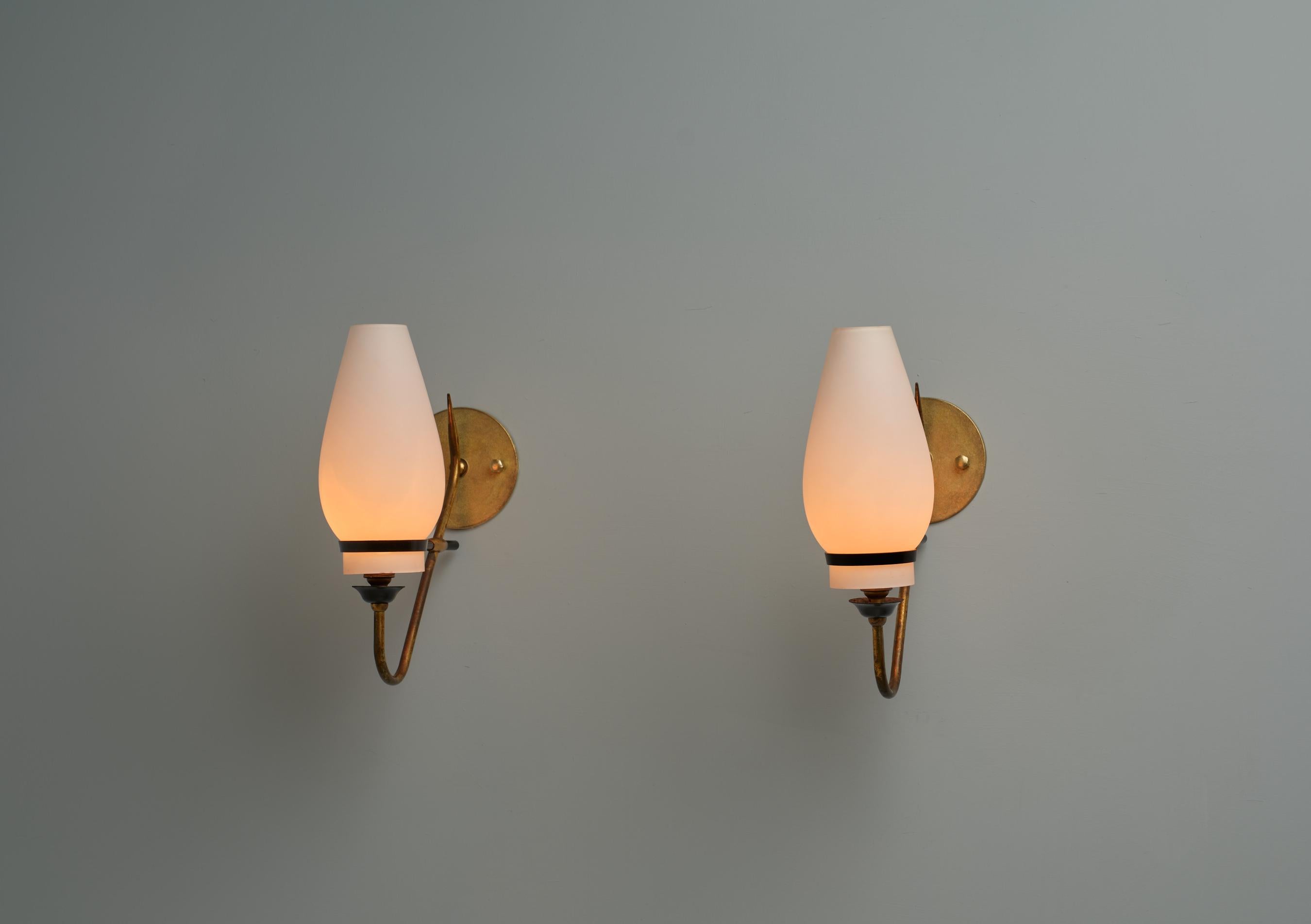 Este par de apliques de pared ejemplifica el diseño italiano de mediados de siglo, elaborado en la década de 1950. Cada candelabro presenta un brazo curvado de latón con una pátina natural envejecida, complementado con una base y un elegante soporte
