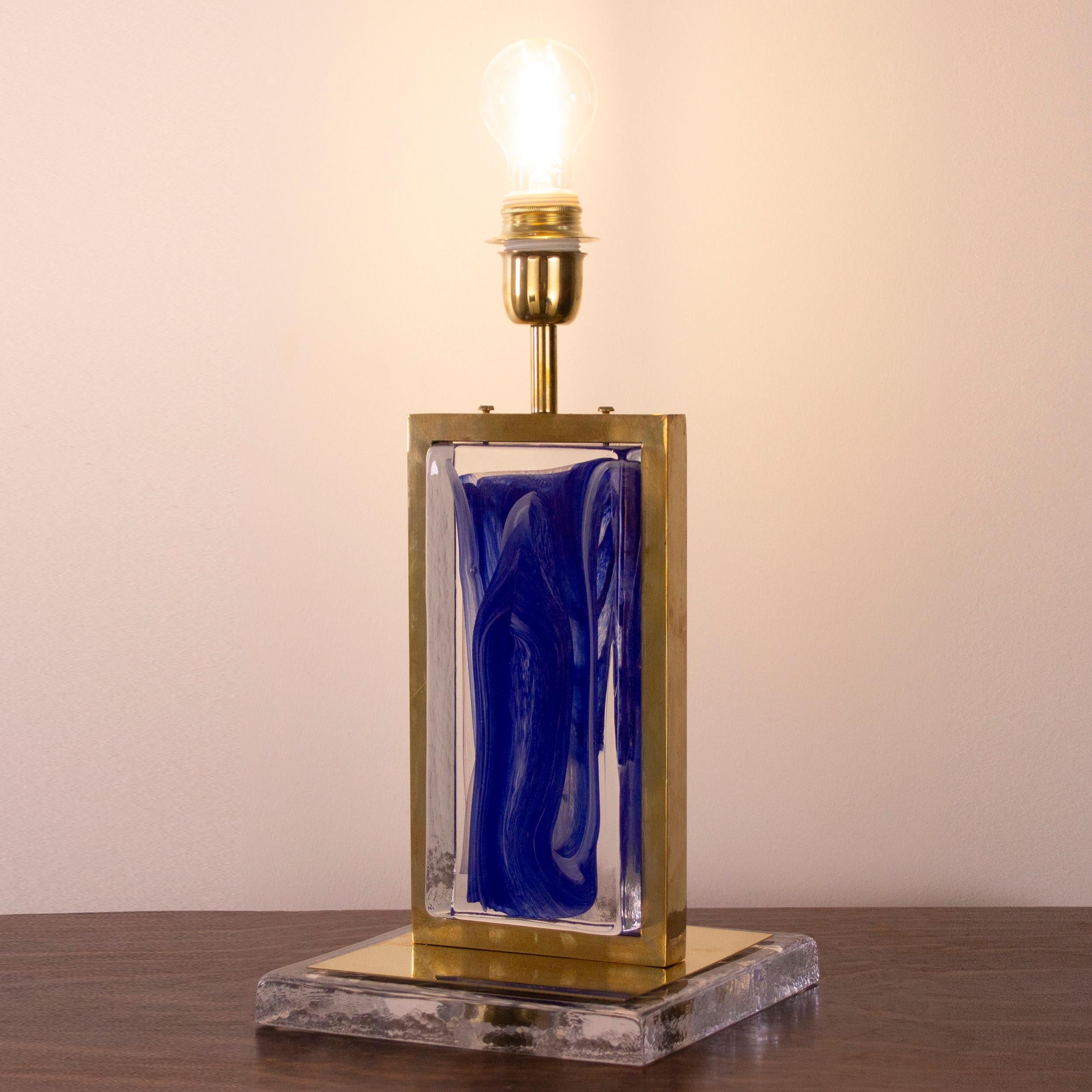 
Una llamativa lámpara de sobremesa hecha a mano en Italia, con una sólida estructura de latón que enmarca un escultural bloque de cristal de Murano. El elemento de cristal muestra un rico tono azul cobalto con fluidos remolinos blancos, creados