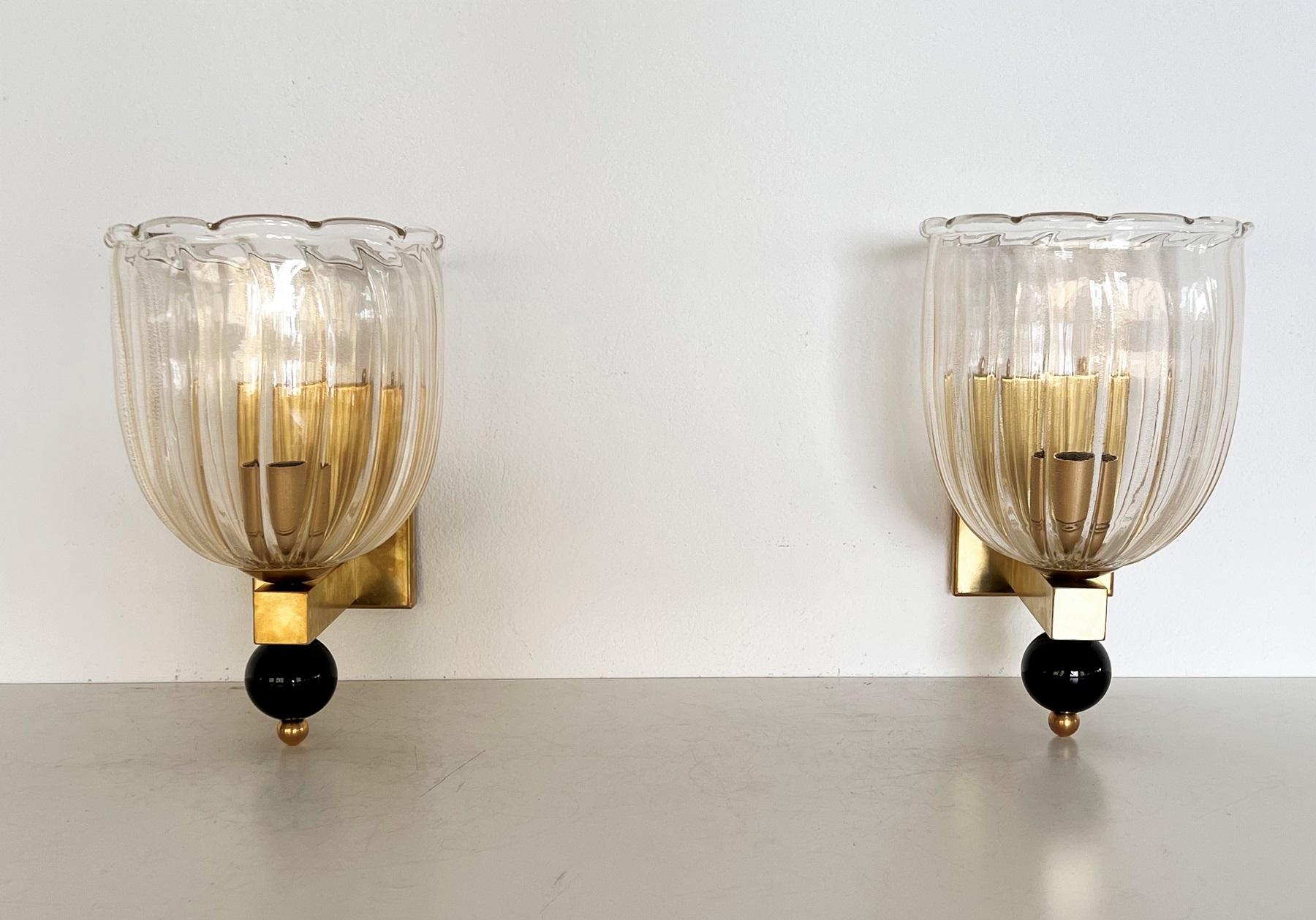 Beautiful set of two gorgeous brass wall lamps made of strong brass base and light transparent Murano glasses with golden shimmer/glitter inside the glass. 
At the bottom of the wall lamps are large glass balls, also handmade from black Murano