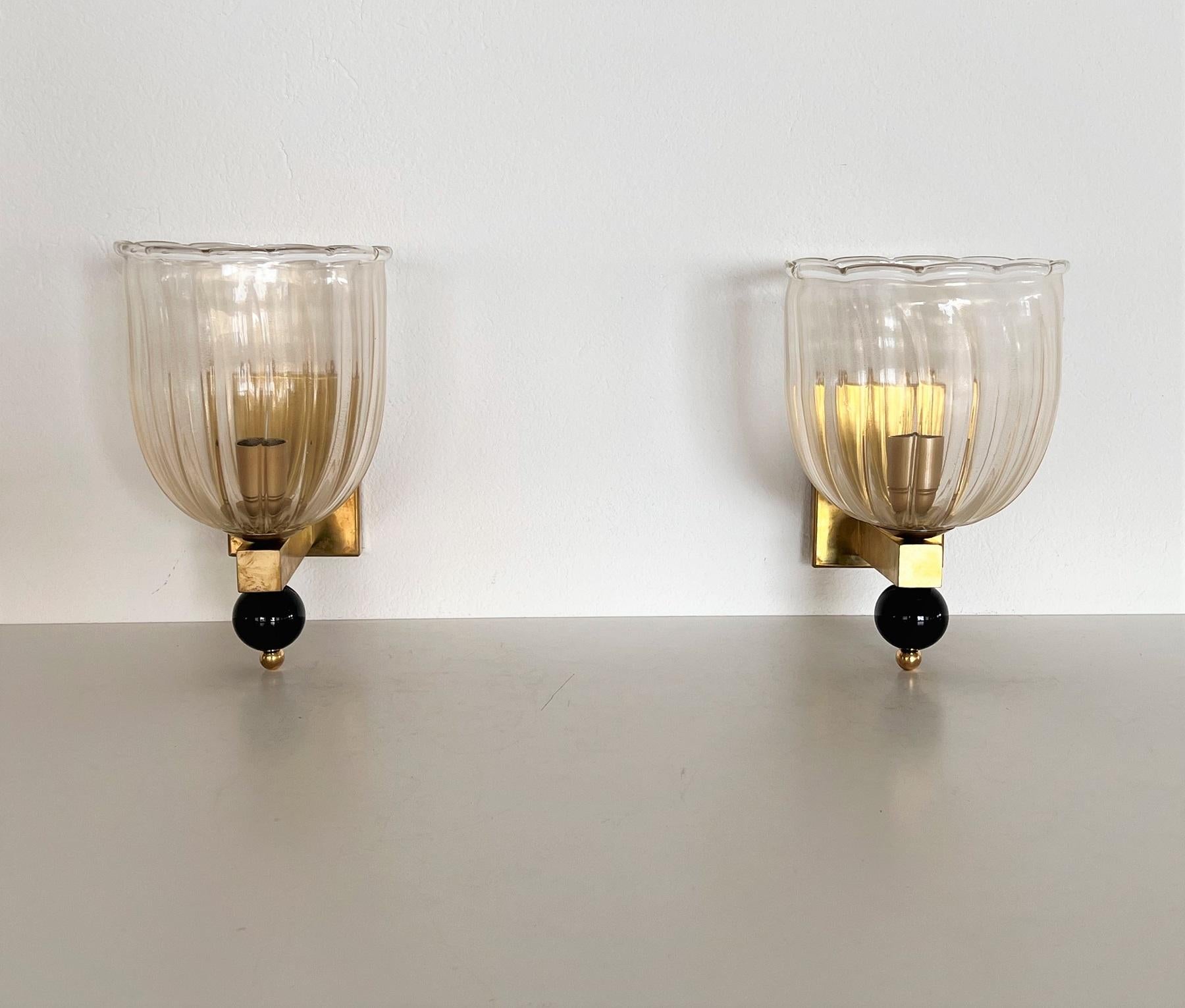 Beautiful set of two gorgeous brass wall lamps made of strong brass base and light transparent Murano glasses with golden shimmer/glitter inside the glass. 
At the bottom of the wall lamps are large glass balls, also handmade from black Murano