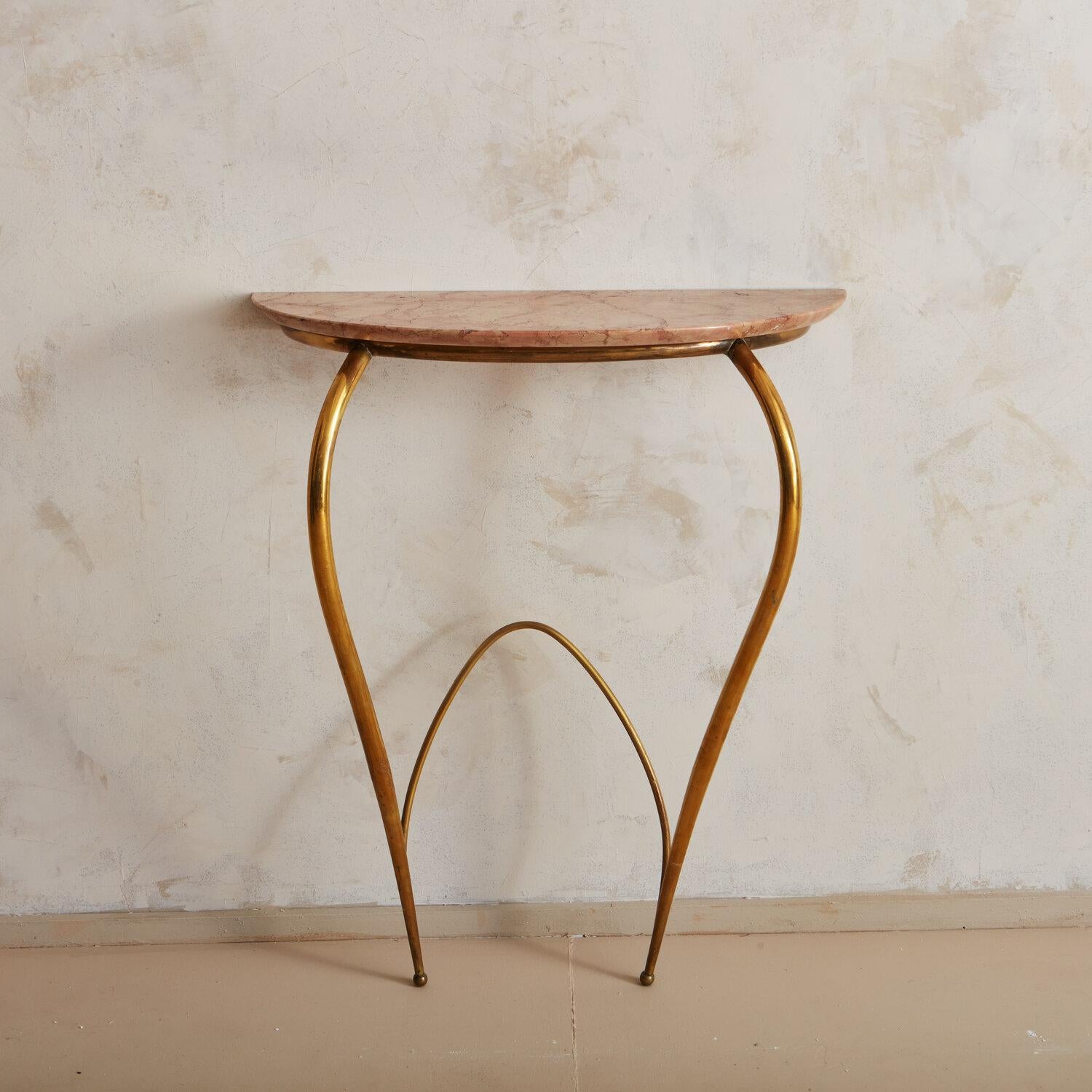A petite console table for a hallway or entryway; this features a curved brass frame and a pink marble demilune shaped top. This table is meant to mount directly to the wall; it is not freestanding.