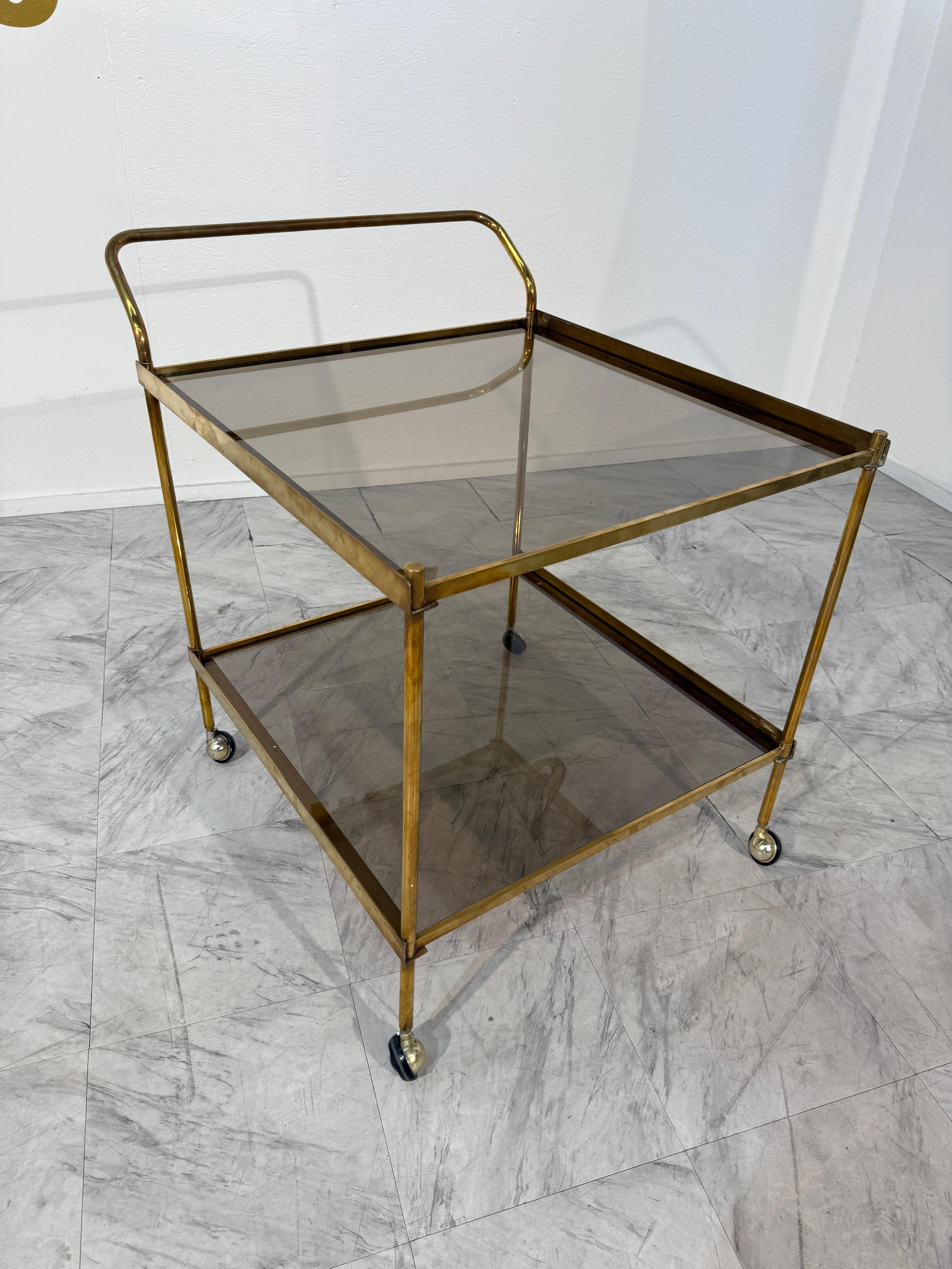 This vintage Italian bar cart features a golden brass frame paired with smoked glass shelves, resting on petite caster wheels that offer both mobility and charm. The clean lines and minimalist structure reflect a distinctly modernist design