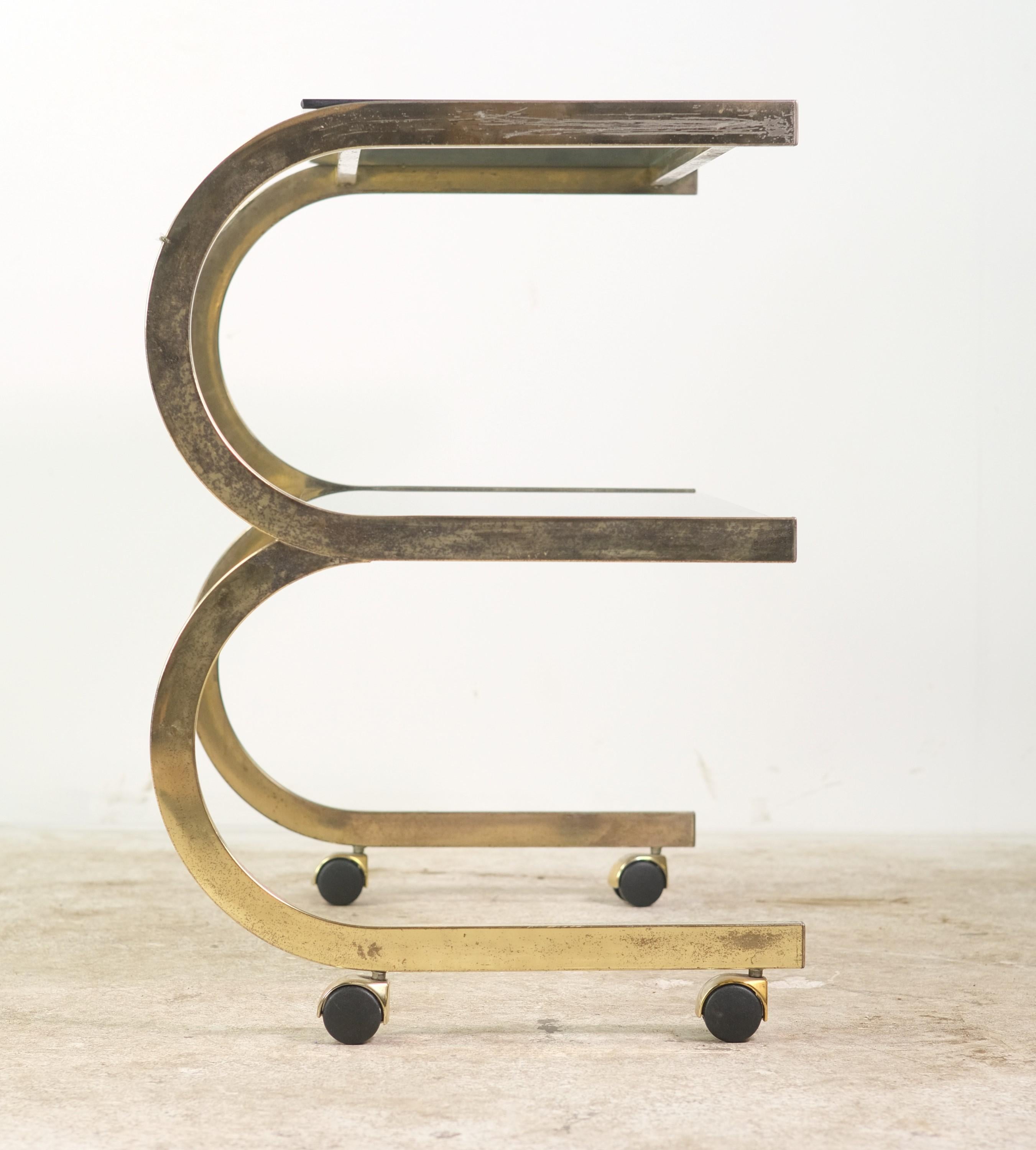 Italian Prandini 2-tier brass bar cart with tinted glass rectangular shelves. This Bauhaus inspired cart has a brass-plated steel frame with wheels. The frame has a looped design supporting the two glass shelves. 

