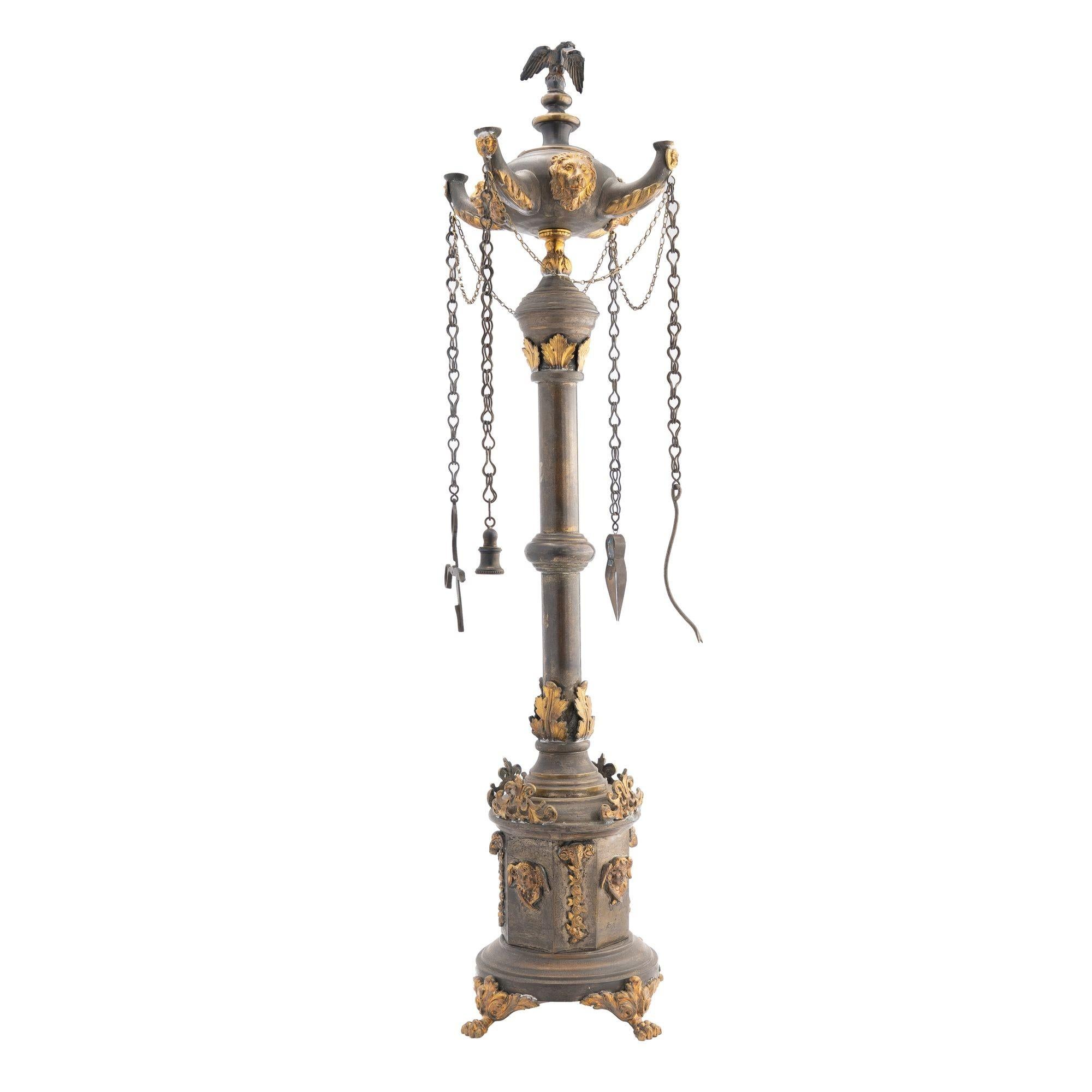 Cast and stamped brass columnar Lucerne lamp with oxidized and contrasting gilt embellishments. The lamp is designed after Roman columnar monument, mounted with a four spout oil font with cover bearing an eagle and four hanging wick implements. The
