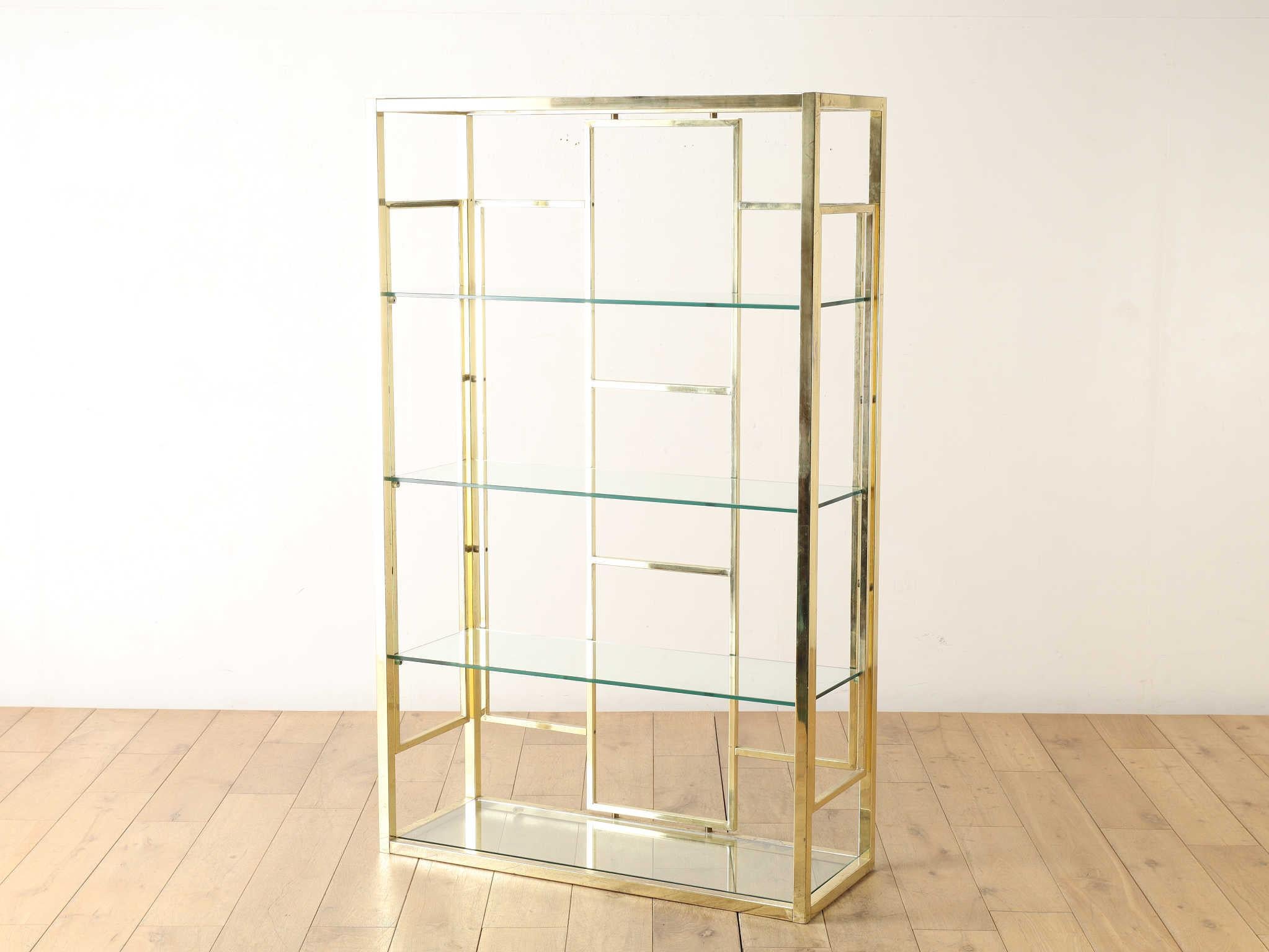Esta estantería vintage italiana, que data aproximadamente de la década de 1970, presenta un llamativo diseño que combina un marco de latón con elegantes estantes de cristal. El refinado contraste entre los cálidos tonos metálicos y la transparencia