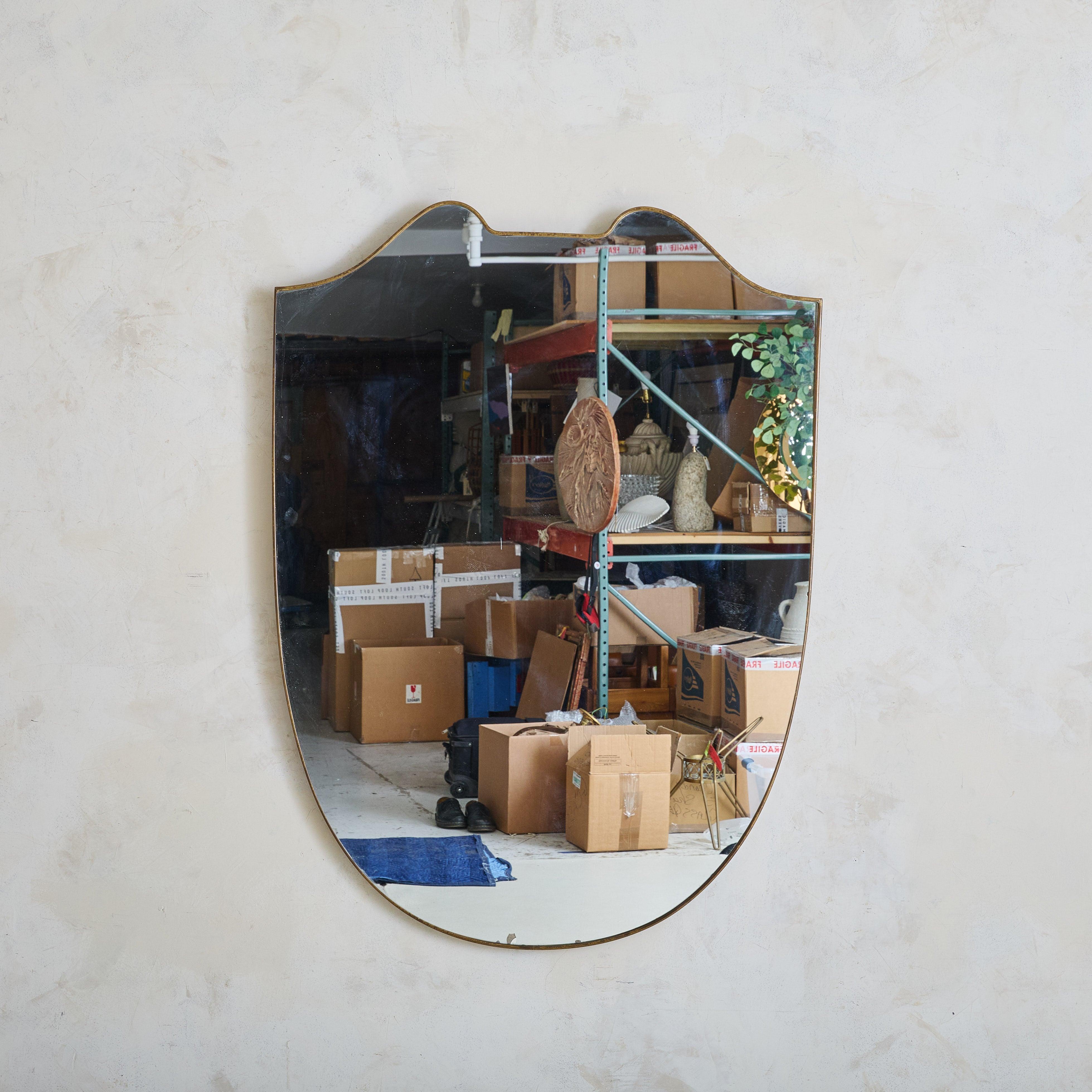Sold and priced individually.

A 1970s Italian shield shaped wall mirror featuring elegant curves along the top and a subtly tapered silhouette. This mirror has a delicate brass frame with a beautiful aged patina. A wooden backing supports the