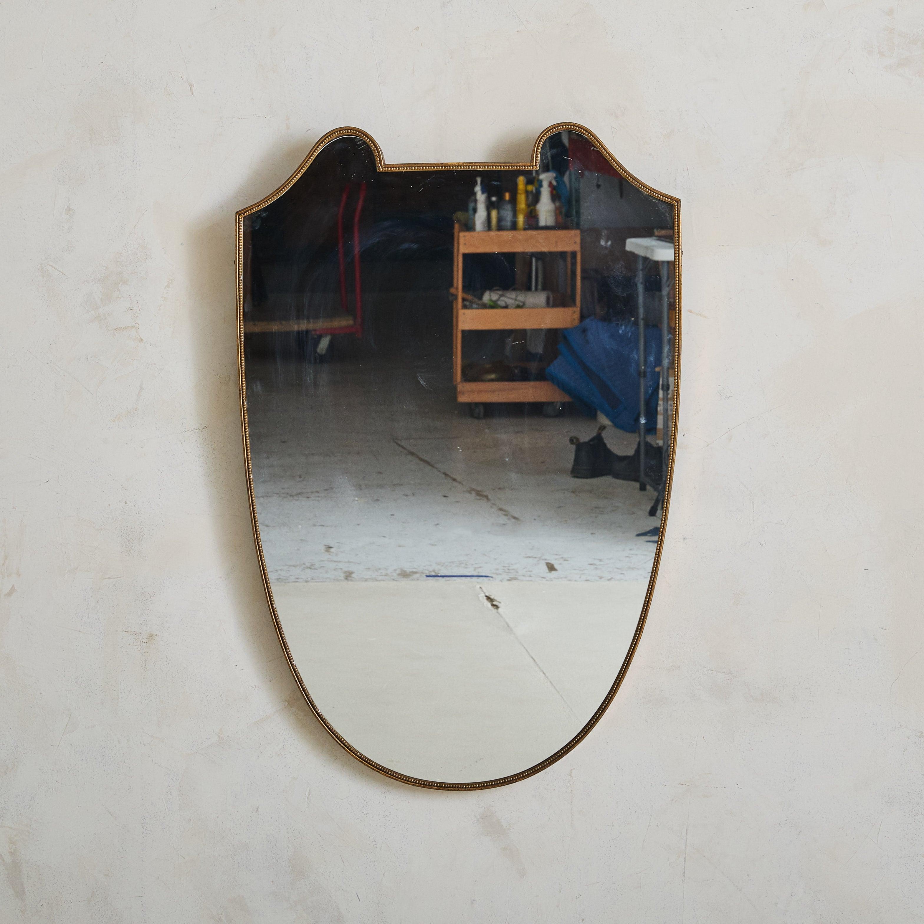 Specchio da parete italiano a forma di scudo degli anni '70, caratterizzato da eleganti curve lungo la parte superiore e da una silhouette sottilmente affusolata. Questo specchio ha una delicata cornice in ottone con una bellissima patina