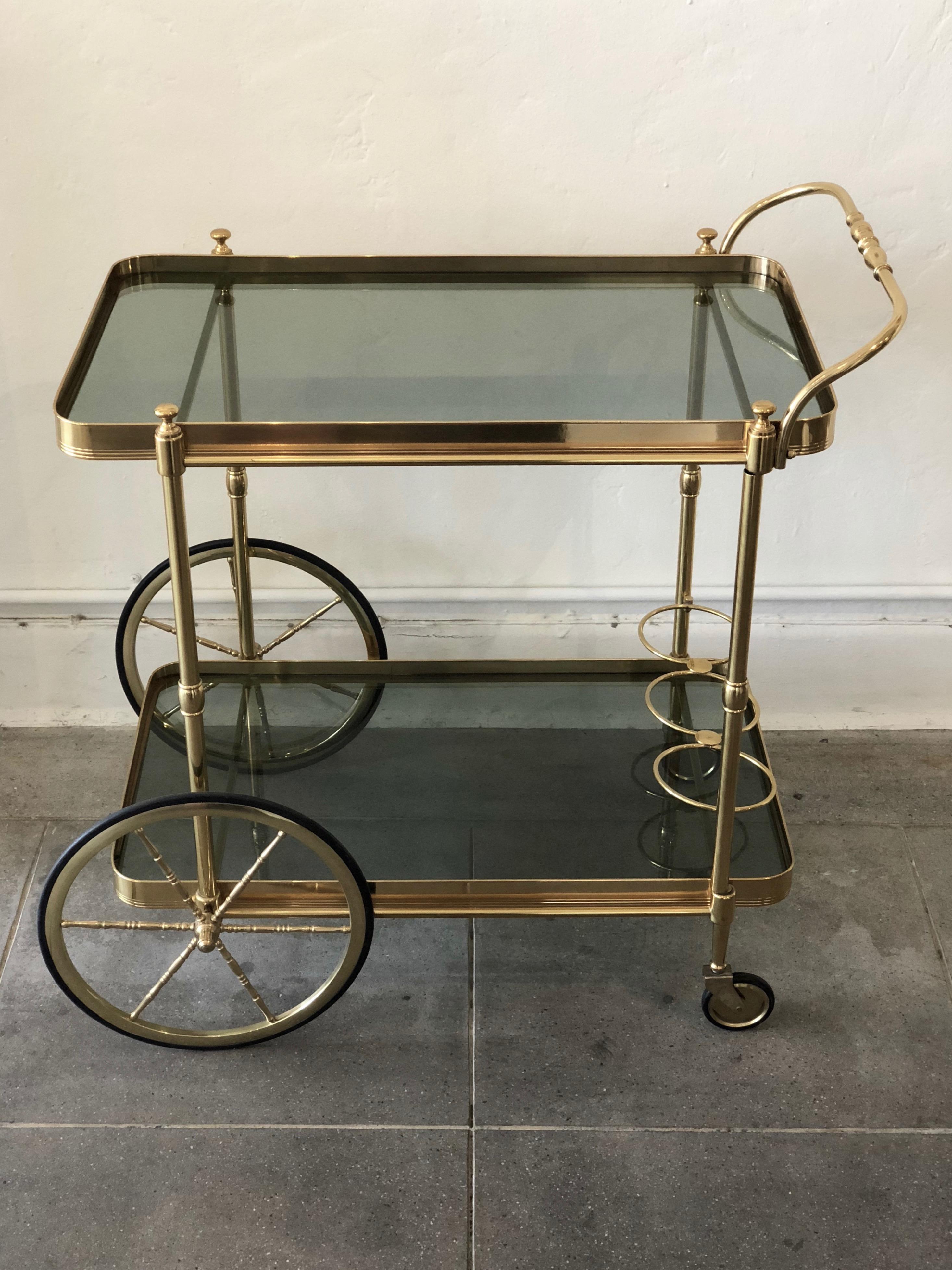 This vintage bar cart is made in Italy and has a nice medium brass tone. There are two tiers of shelving with smokey grey colored glass. The bottom shelf has three bottle spaces. The wheels and casters are in working order. 
The measurements below