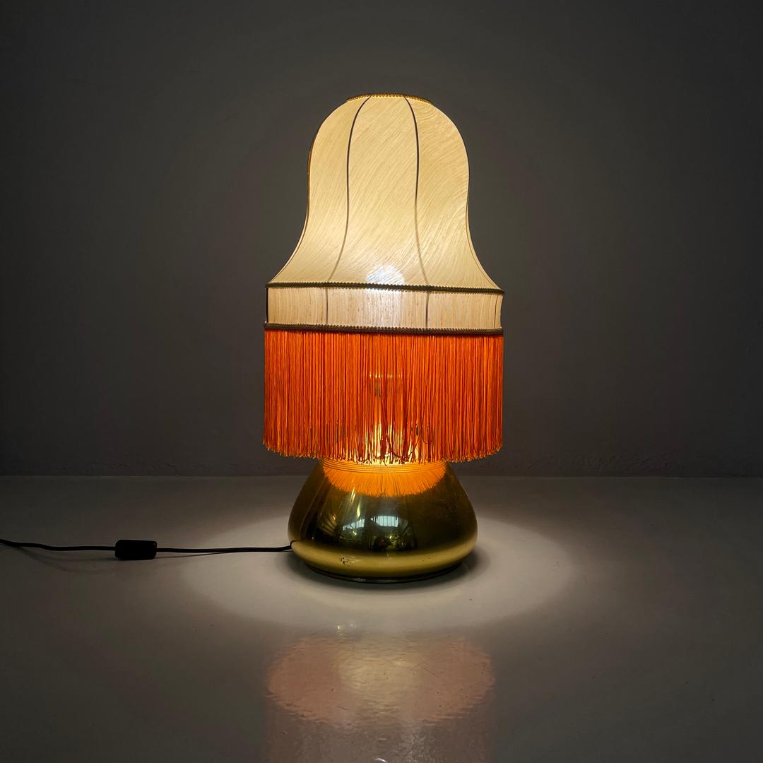 Italian brass table lamp with beige lampshade and orange fringes, 1980s
Round base table lamp. The lampshade is round in beige fabric, and follows sinuous curved shapes that start from the top and widen in the center. It ends at the bottom with a