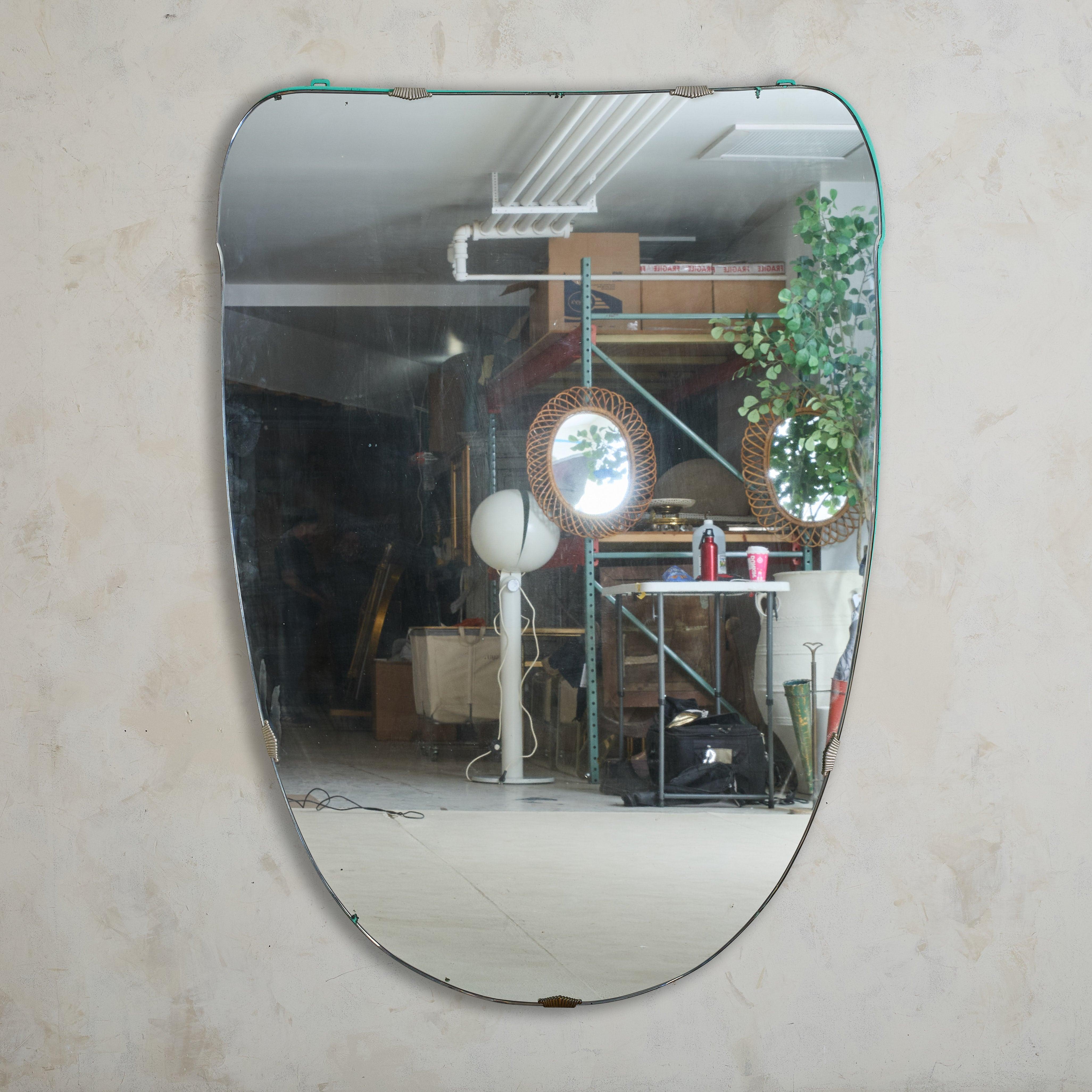 Ce grand miroir mural italien des années 1970 présente une forme doucement arrondie encadrée de laiton, accentuée par de subtiles bordures de bois peintes en turquoise et de délicats détails métalliques festonnés. Vieilli à la perfection, le verre