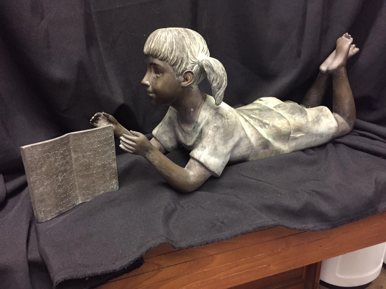 Cute Italian bronze figure of a pigtailed little girl laying on her belly with her legs crossed enjoying a book. Weathered patina. 
Early 20th century.