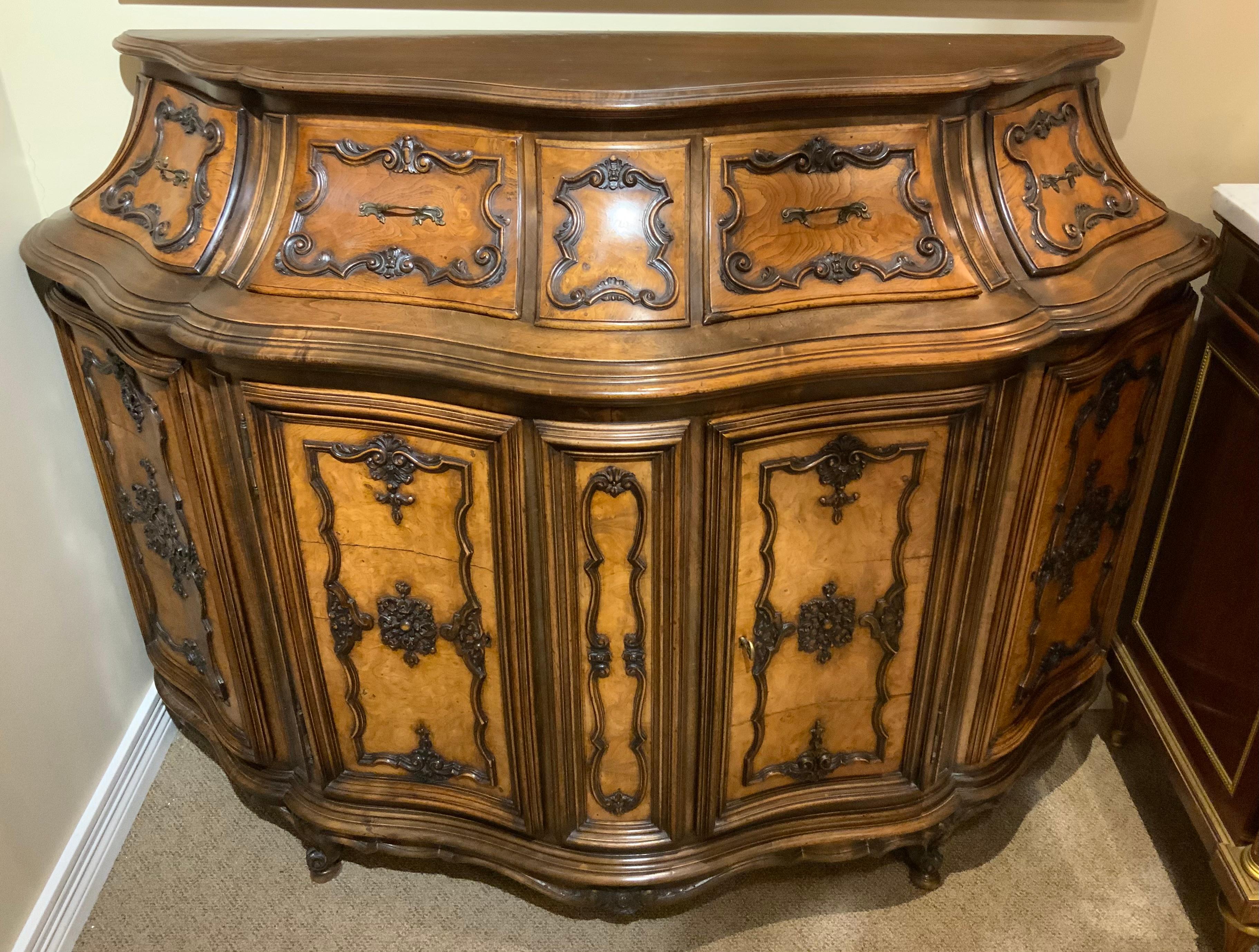 The serpentine shape of this piece makes it a real statement 
It is made of a burr walnut in a honey hue.  It has two doors
That open to a shelved storage. It has two drawers that
Open easily. It has finely shaped decorative wood mounts
The side