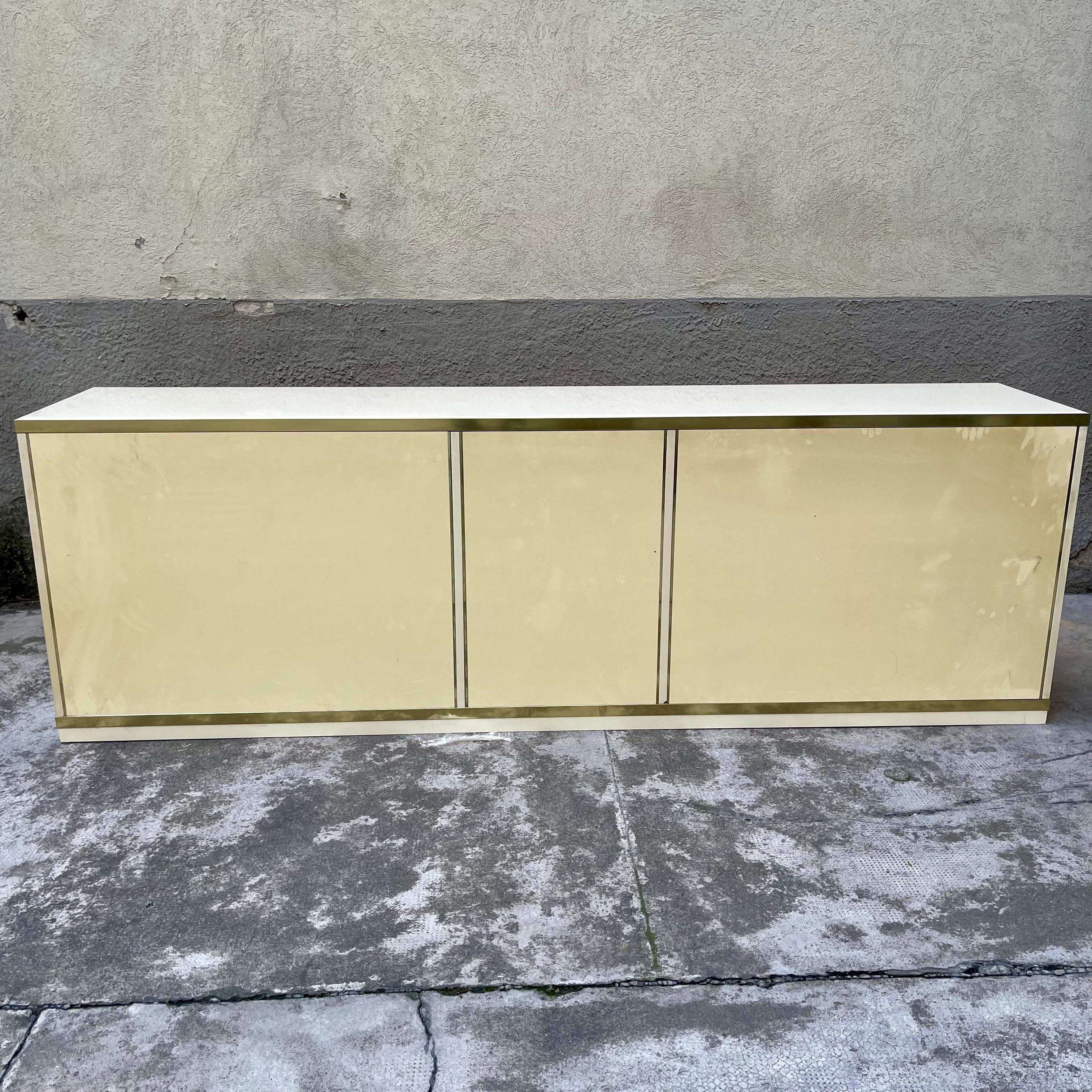 This sideboard, constructed of ivory-colored laminate and embellished with brass beading and detailing, has push-to-open doors and drawers. It is part of a complete environment, produced by the company Sabot.
The cabinet is combined with a table and