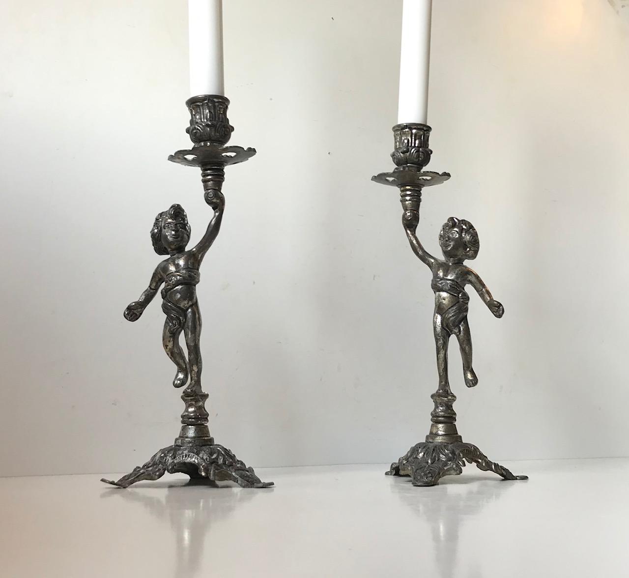 A pair of metal-alloy candlesticks with cherubs. Each cherubs with different facial expression. Manufactured in Italy during the 1940s or 1950s. Both of them stamped: Made in Italy beneath their base. This set uses regular sized candles.