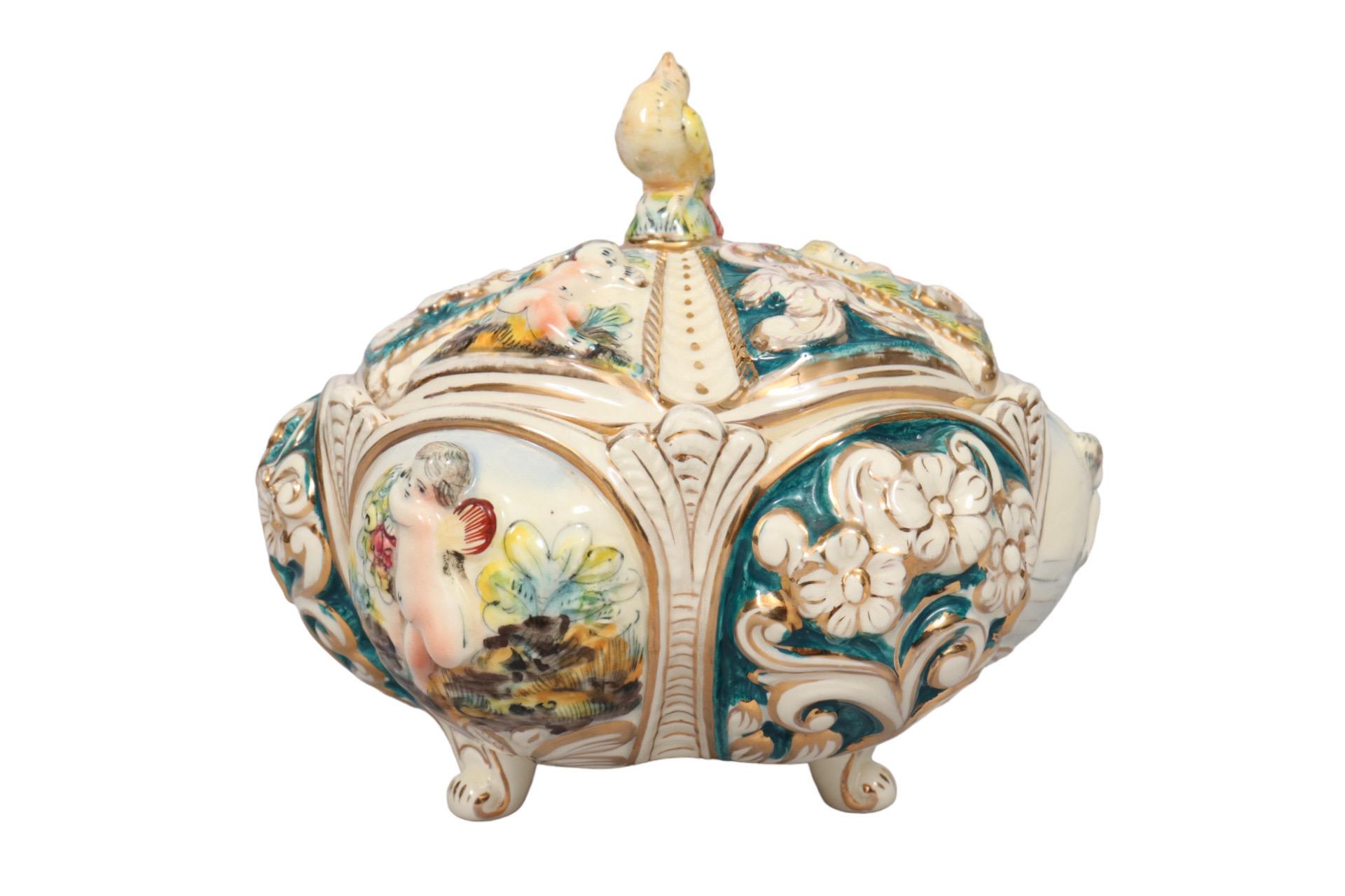 An Italian Capodimonte style ceramic lidded bowl. Decorated with six faces in a bombé shape. Playful embossed cherubs alternate with embossed flowers painted in lavish jewel tones edged in gold. The lid is similarly decorated, topped with a colorful