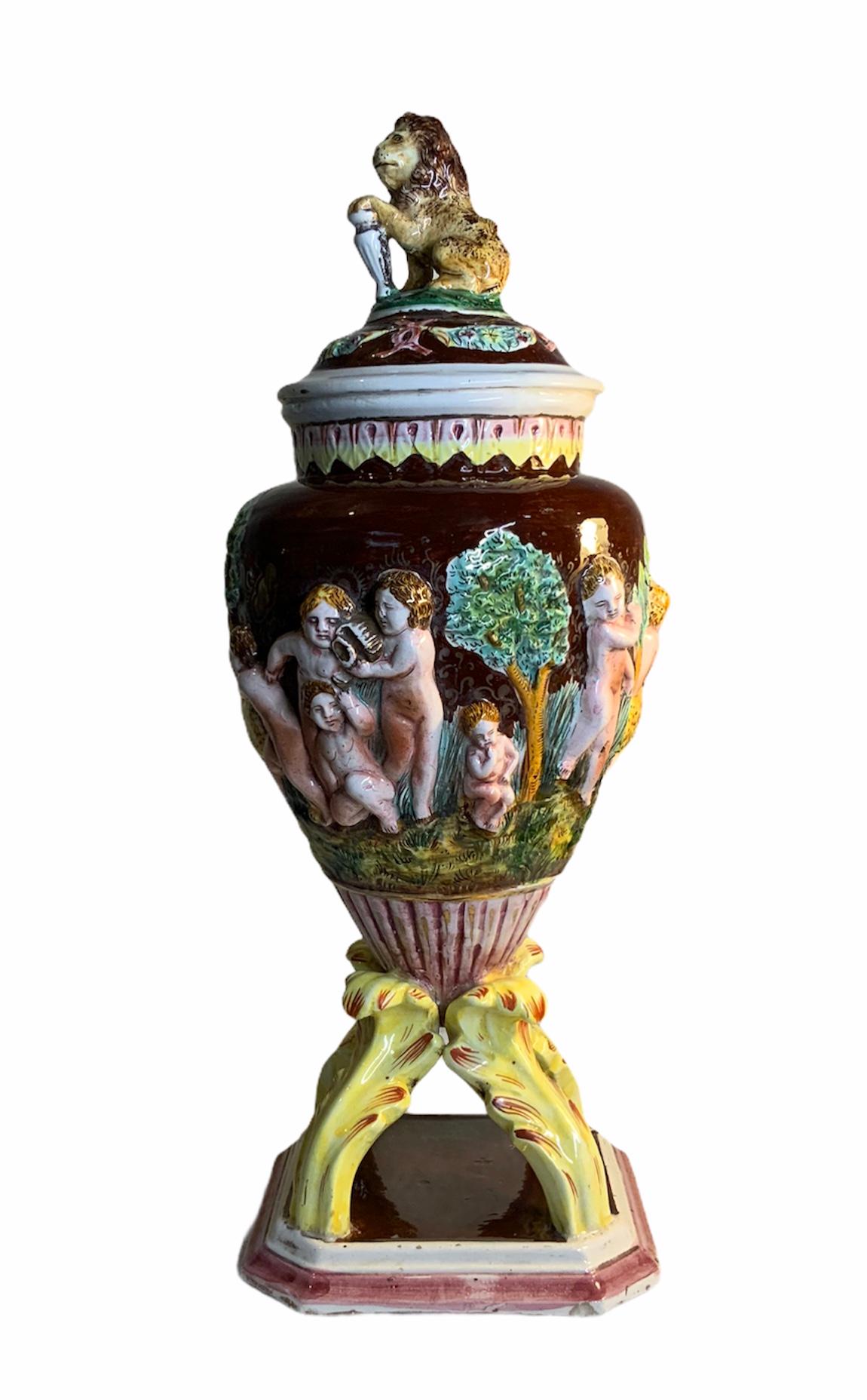 This is an Italian hand painted porcelain urn with a wine color background adorned with a continuous scene of a repousse group of nudes cherubs in the jungle. Some of them are playing with a tiger and others are wondering around. The urn is