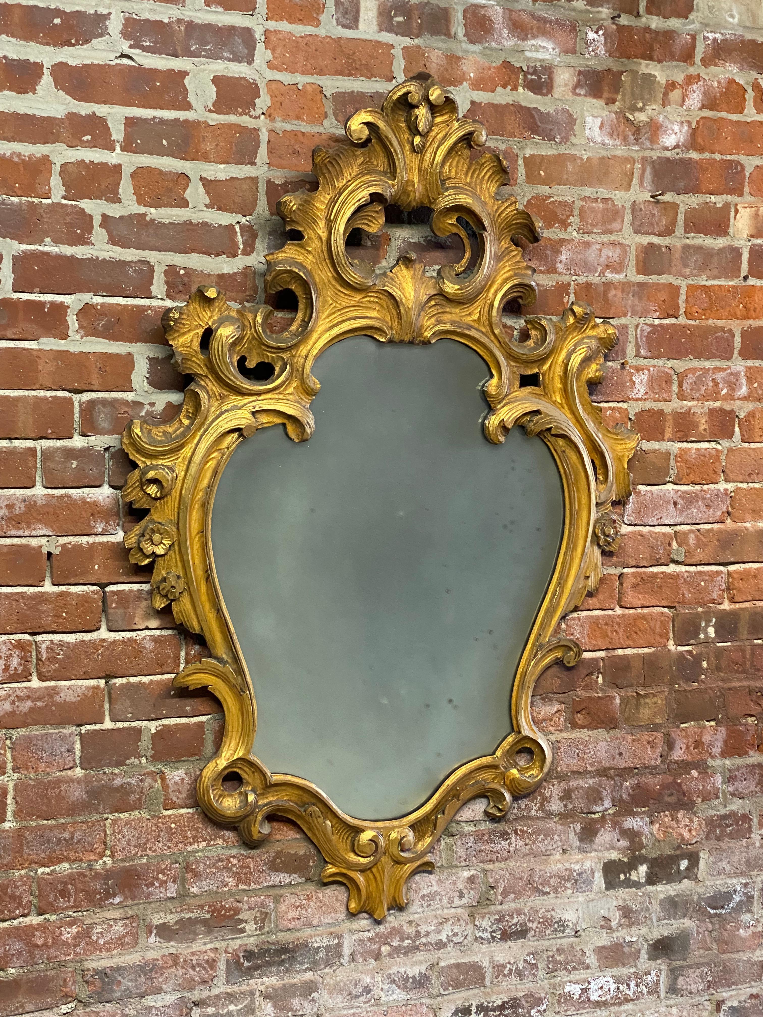Italian carved and gilded scroll and floral wall mirror. Hand carved and gilded wood frame. Red bole and gesso substrate. circa 1940-50. Good overall condition with wear consistent with age and use. Some minor losses and chips to the wood and