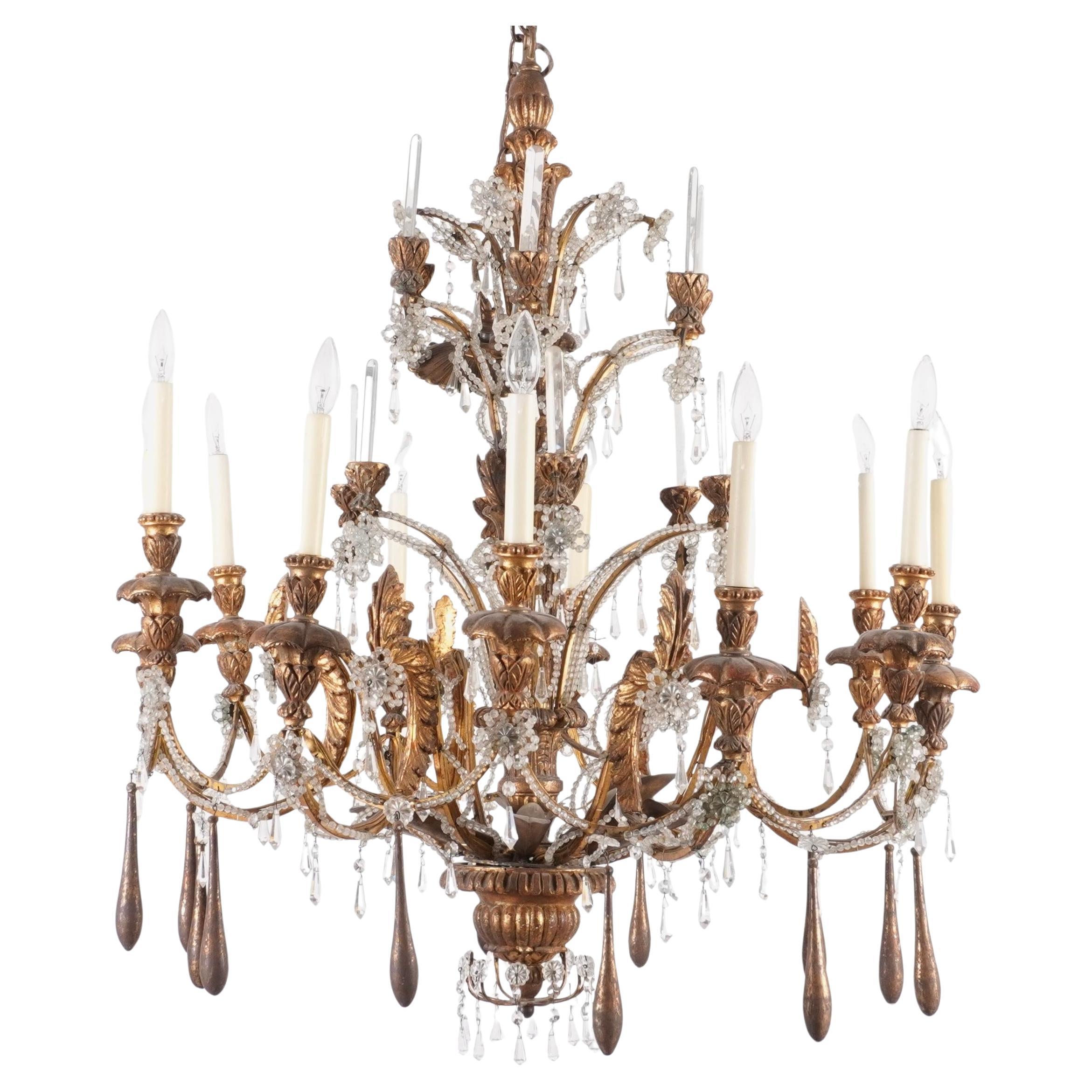 Lustre italien à deux niveaux en bois doré sculpté, fer et cristal perlé. Ce lustre en bois doré sculpté à la main est orné de cristaux et de perles de cristal taillés à la main, ce qui lui confère une élégance d'antan. Avec ses douze bras en