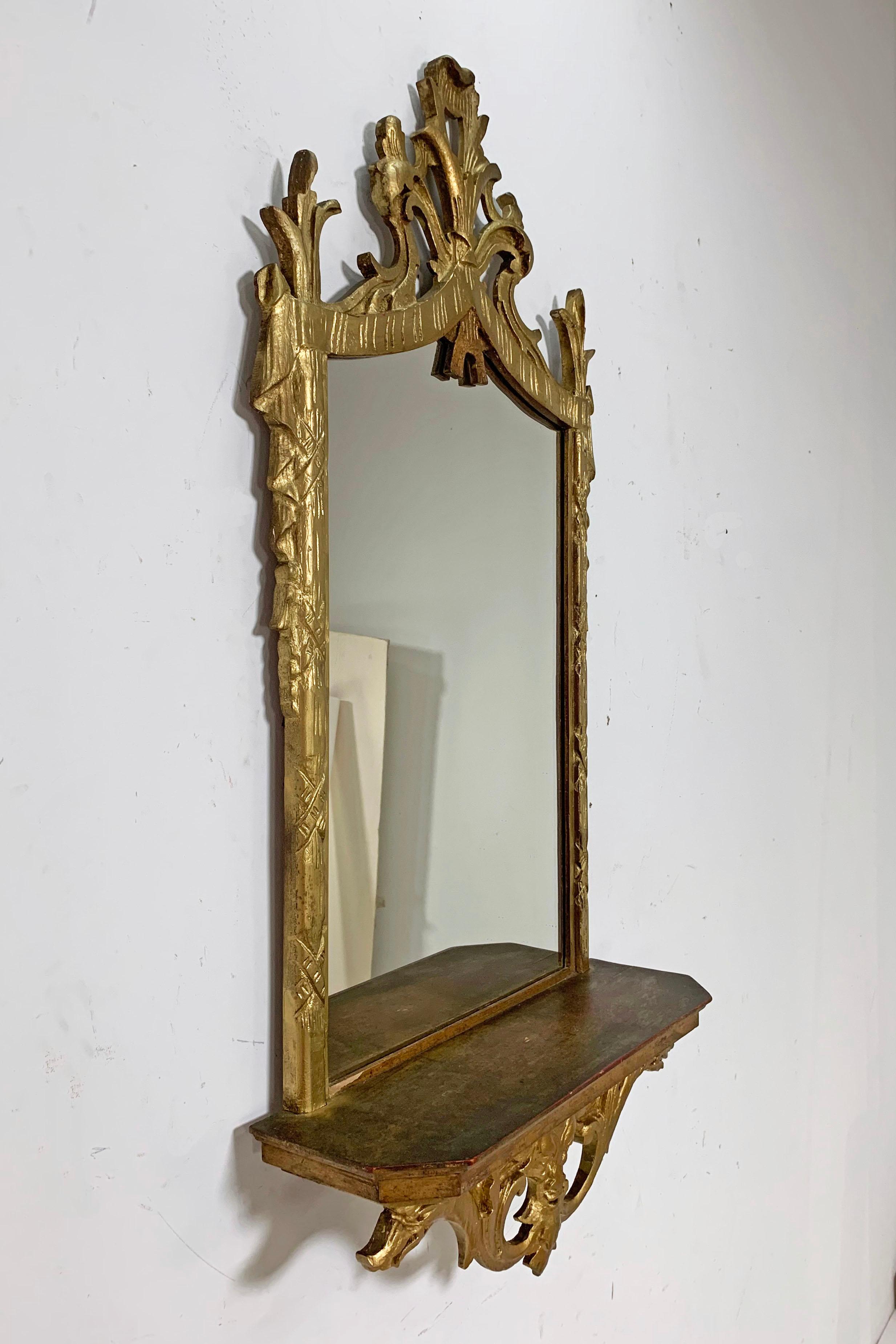 Italian carved giltwood mirror with under shelf, circa 1950s. Elaborate top garland carvings with side pedestals of Roman fasces, and images of Poseidon and dolphins to lower crest.