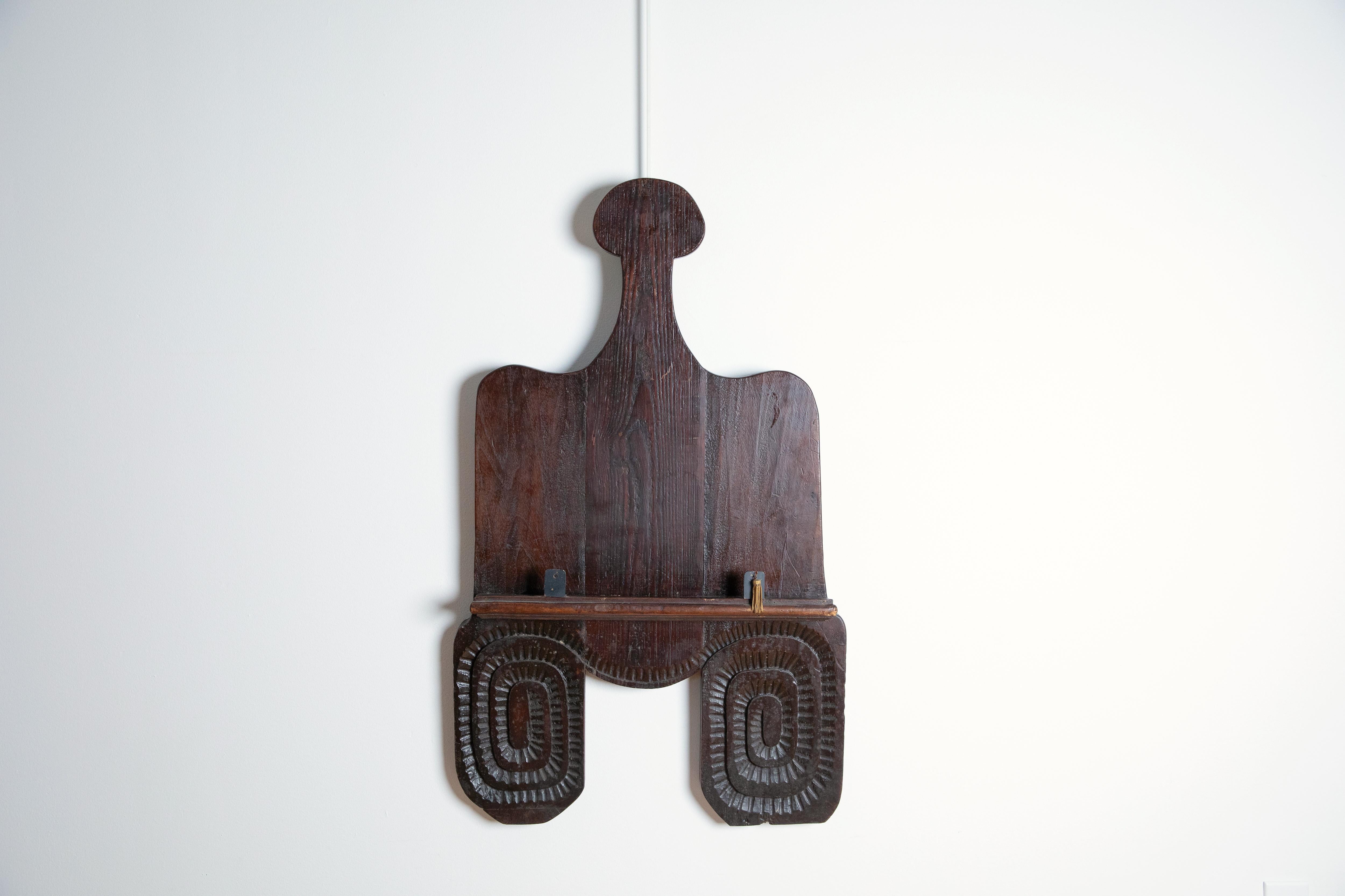 in the shape of a bread board with rounded top corners this wall hanging features a ledge for a book or artwork, the bottom double terminals guilloche carved