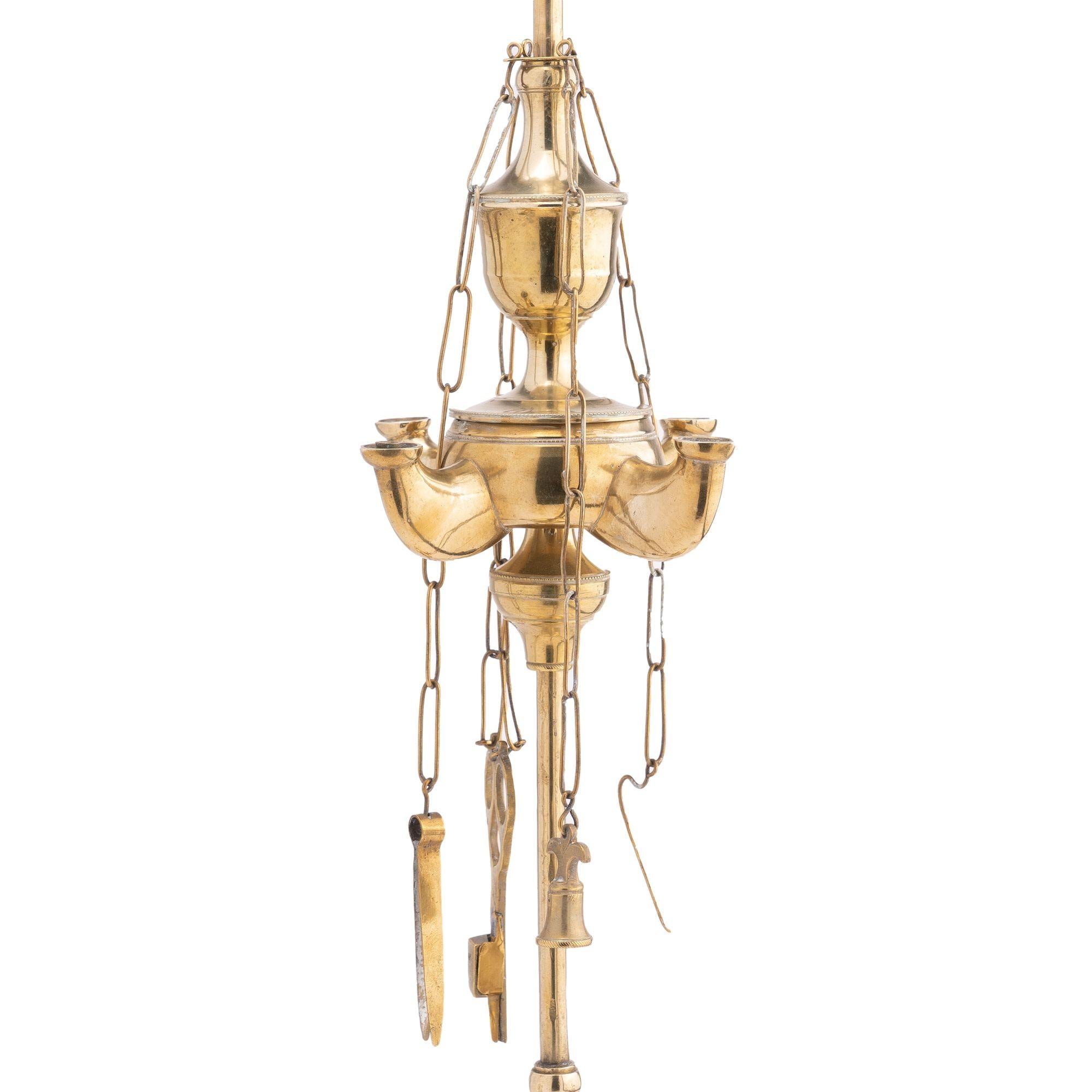 Cast brass four burner Lucerne oil lamp on a cast threaded rod. The rod is topped with an open shield form finial and is treaded to a conical centered circular base with standing rim, drip pan. The font is supported by an adjustable sliding knob and