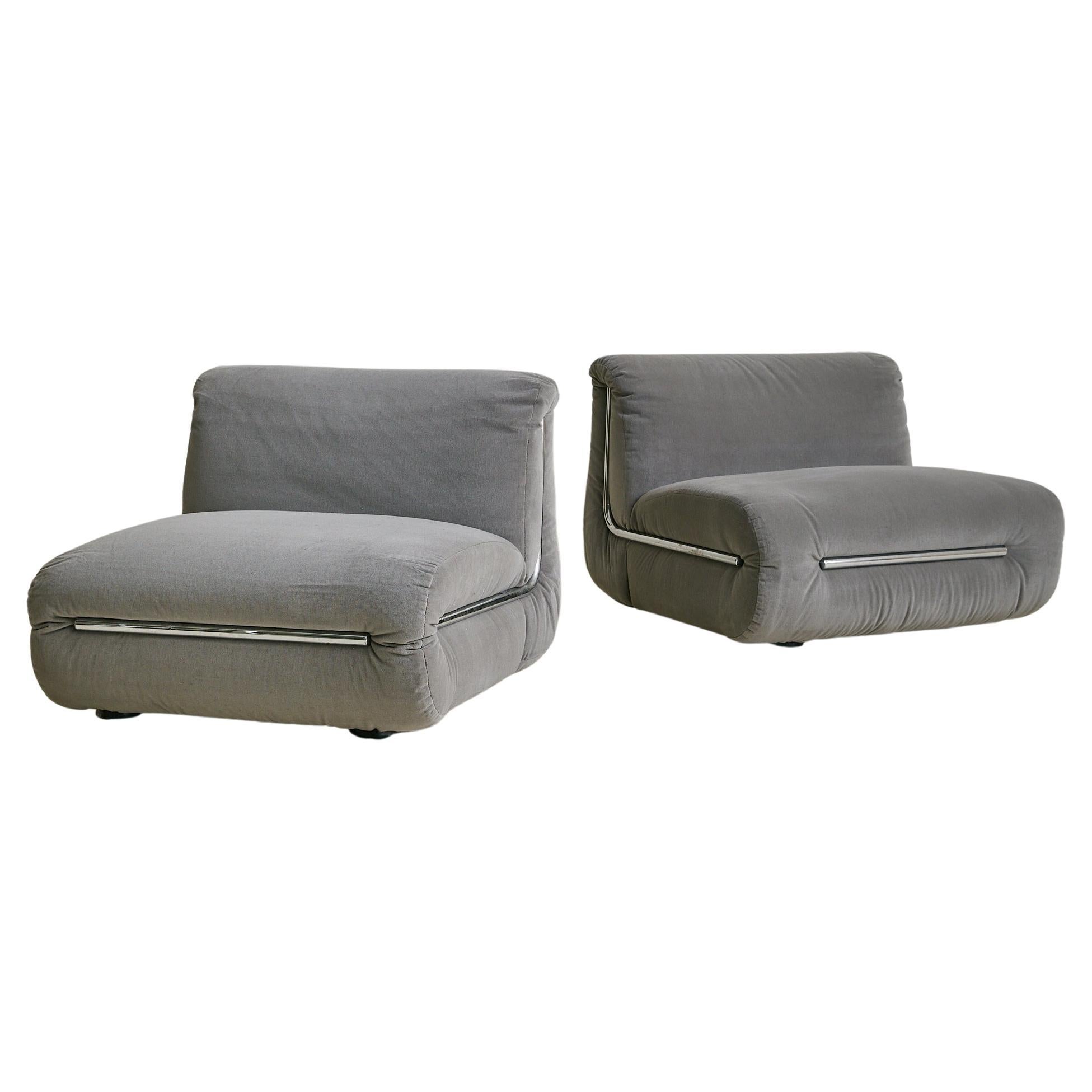 A 1970s Italian lounge chair in dove gray velvet, with a low, generous seat and gently curved back. The form is minimalist and rectilinear, softened by rounded edges and deeply padded seat and back cushions. Chromed steel tubes wrap along the sides