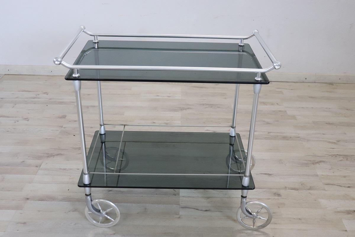 te ofrecemos este refinado carrito de bar fabricado en Italia en torno a los años 80. Se caracteriza por una estructura de metal cromado y dos estantes de cristal. En la parte inferior, un separador en la parte superior permite guardar botellas. Las
