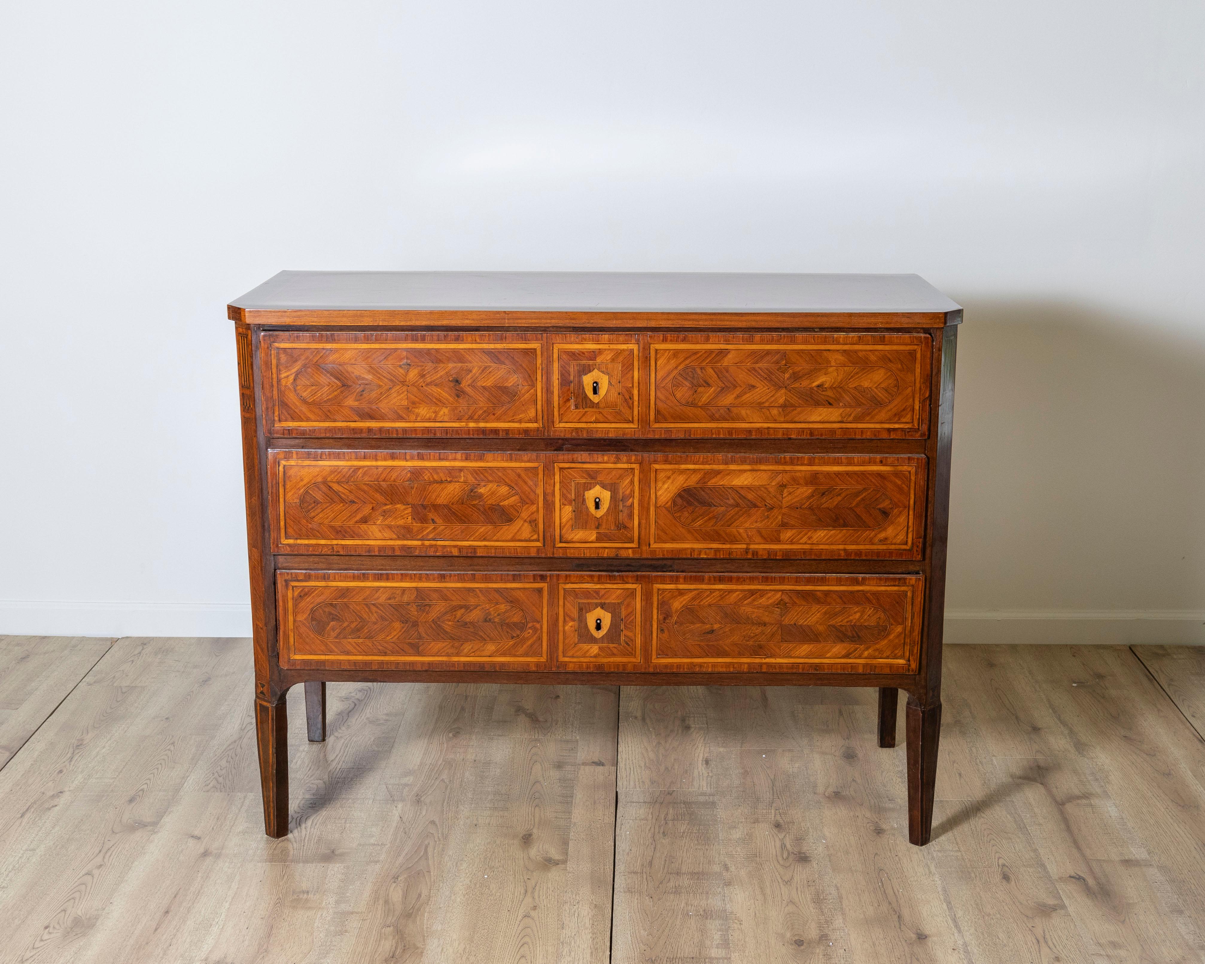 Une exquise commode italienne du XVIIIe siècle, à la marqueterie magistrale et à l'élégance intemporelle. Cette pièce raffinée comporte trois longs tiroirs, chacun orné de motifs géométriques incrustés dans des placages de bois clair et foncé
