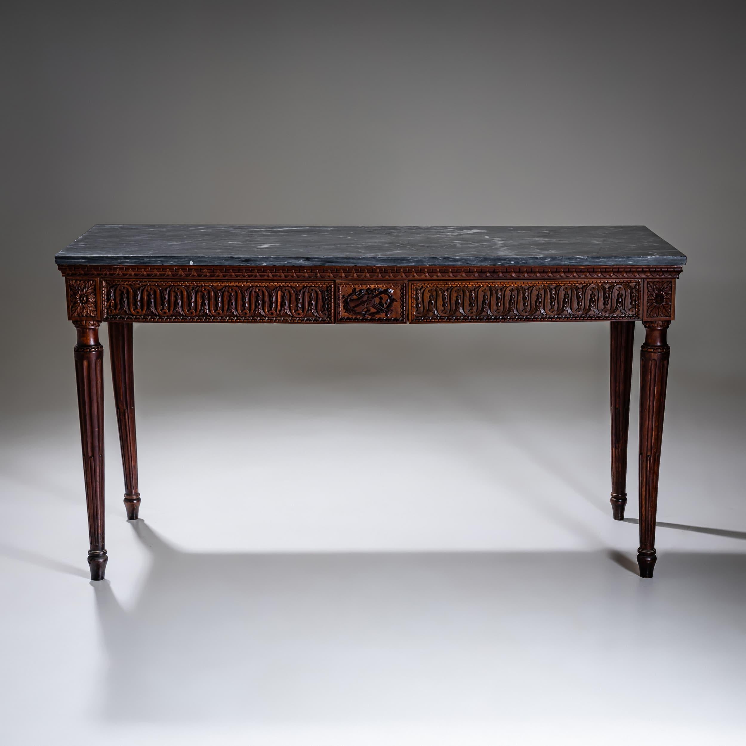 A refined Italian console table dating to the late 18th century. The piece stands on four tapered and fluted legs and features a richly carved apron adorned with stylized leaf motifs, rosettes, and classical ornaments. The front is highlighted by a