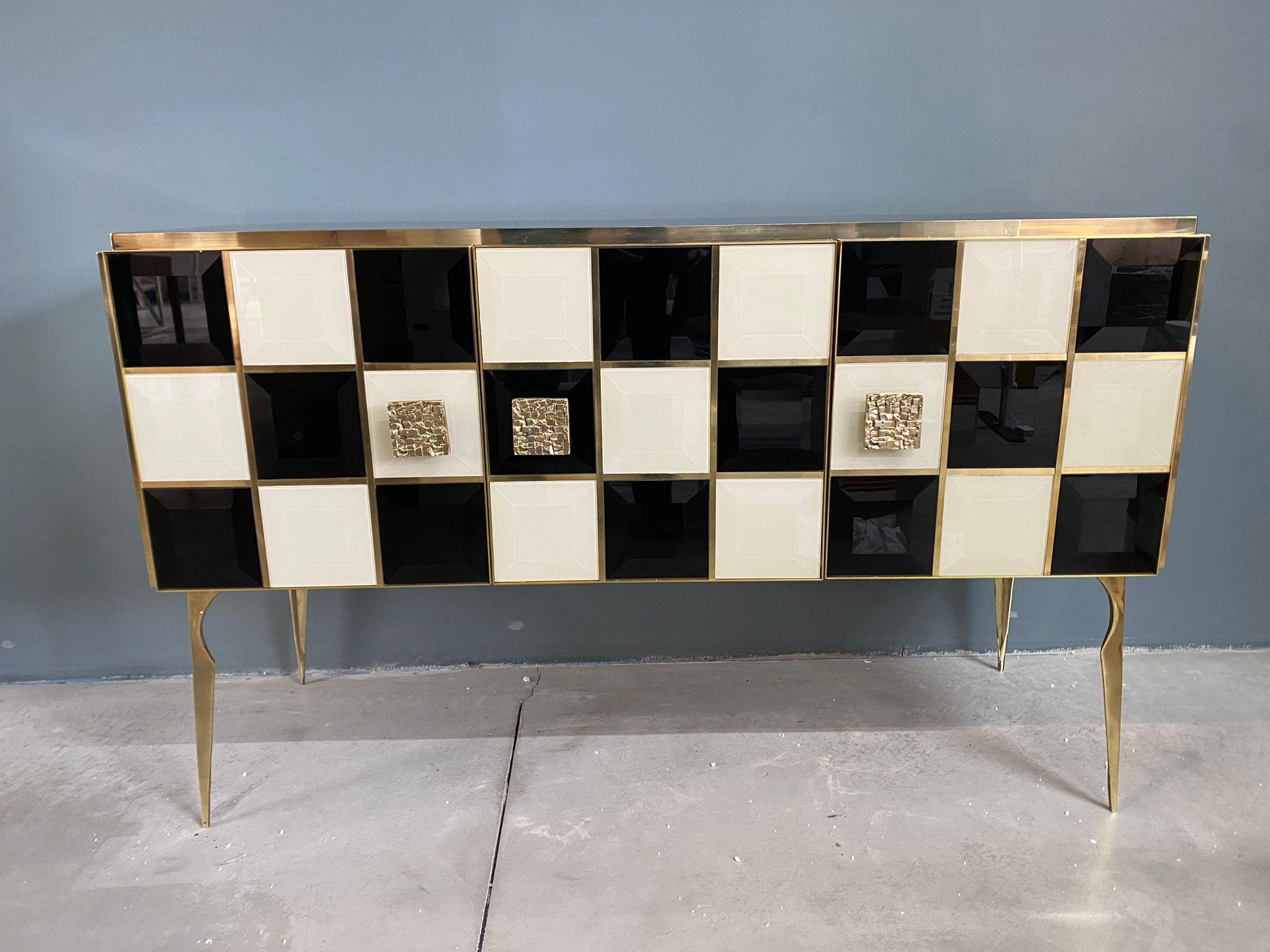 Precioso aparador diseñado y fabricado a mano por nosotros con estructura e interior en madera maciza, patas moldeadas y acabados en latón y tapas y decoraciones en cristal de Murano coloreado en blanco y negro. 
La particular preciosidad de este