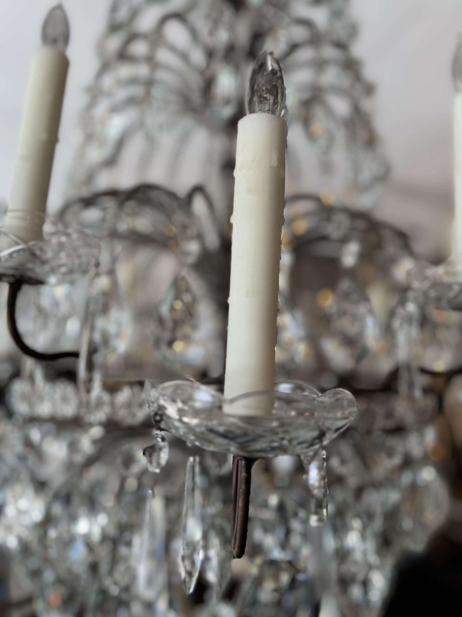Questo lampadario emana un'eleganza senza tempo e una presenza scenica. Progettato con una silhouette a livelli, presenta una splendida struttura in metallo con perline, ornata da gocce di cristallo e rosette che scendono a cascata. 12 bracci a