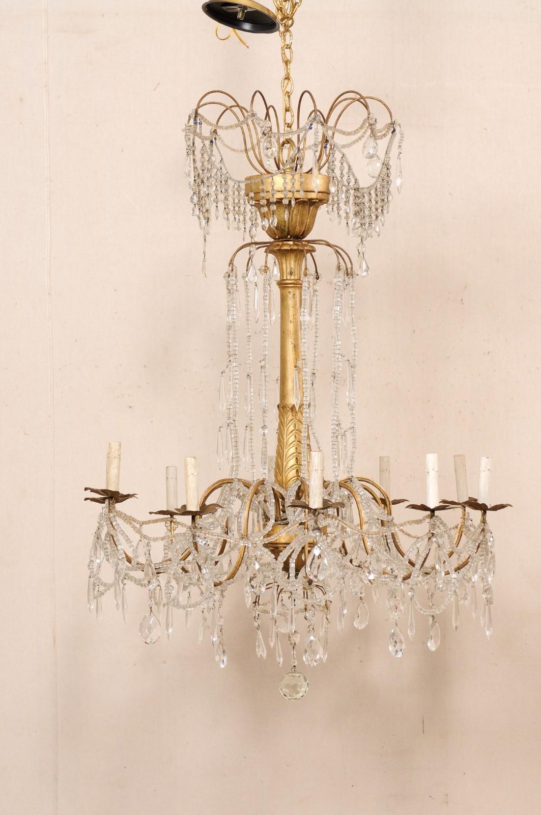 An Italian crystal and gilt wood column chandelier from the mid 20th century. This mid-century hanging light from Italy features a gilt-wood central column, adorn with leaved-wraps carved about it's lower gallery and floral wraps about the top and