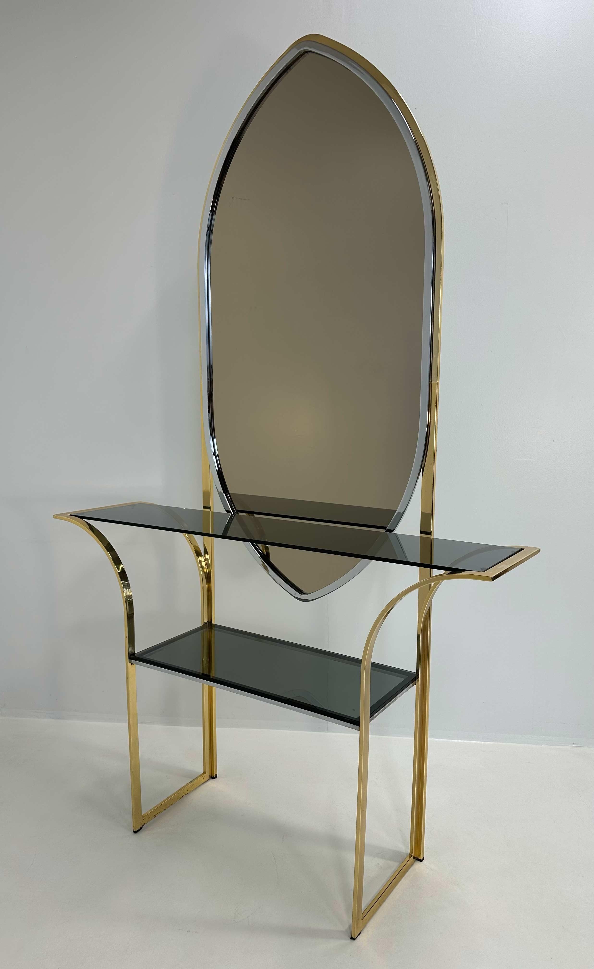 Italian console from the 1970s in brass with smoked glass shelves and a matching mirror. The structure features sinuous lines with curved legs supporting two dark glass shelves. The tall, pointed mirror is framed in chromed metal and integrated into